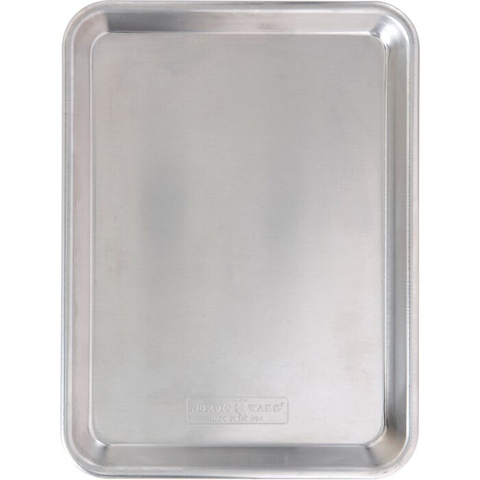 Bageplade i blankt ubehandlet aluminium set skråt oppefra, med ombukket kant hele vejen rundt og et ovalt Nordic Ware-mærke præget i bunden, på hvid bund