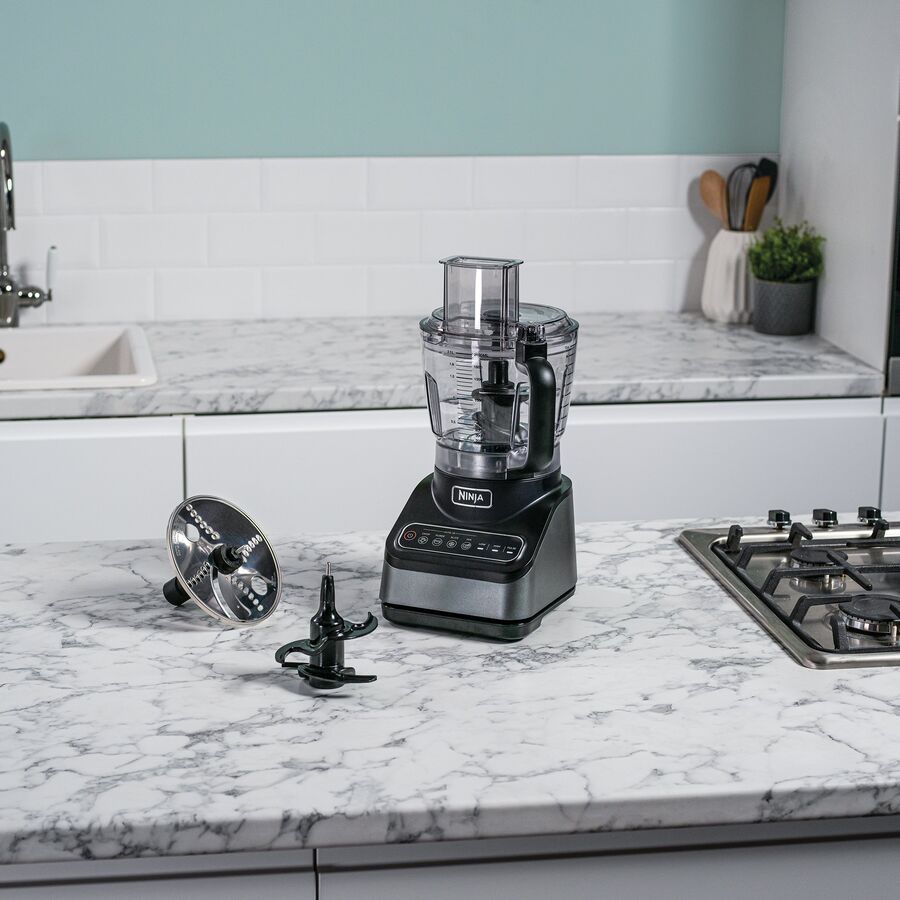 Sort foodprocessor på en hvid marmorbordplade foran en lys grønblå flisevæg, med en riveskive og en kniv liggende på bordet foran den og et gasblus til højre