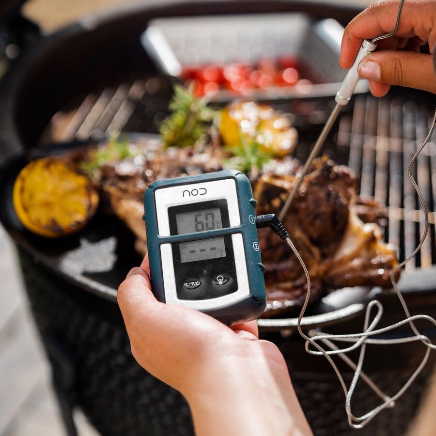 En hånd holder en trådløs modtager med kernetemperaturen foran en kuglegrill med kød og grøntsager