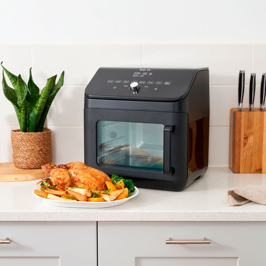 Sort airfryer-ovn med glaslåge på en lys køkkenbordplade foran hvide fliser, med en stueplante til venstre, en knivblok i træ til højre og et fad med brunet kylling og ovnbagte grøntsager foran