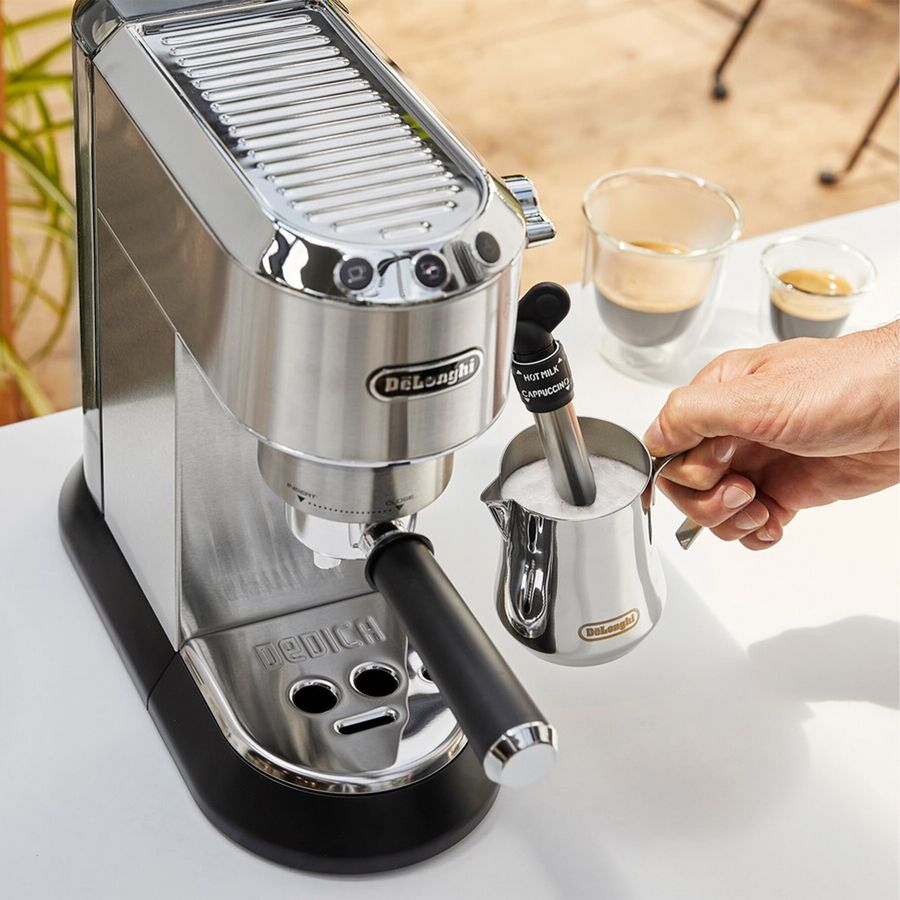 Espressomaskine i børstet stål på en hvid bordplade, hvor en hånd holder en lille stålkande op under dampdysen, med to glas espresso bag maskinen og en grøn plante og et varmt gulv ude af fokus bagved