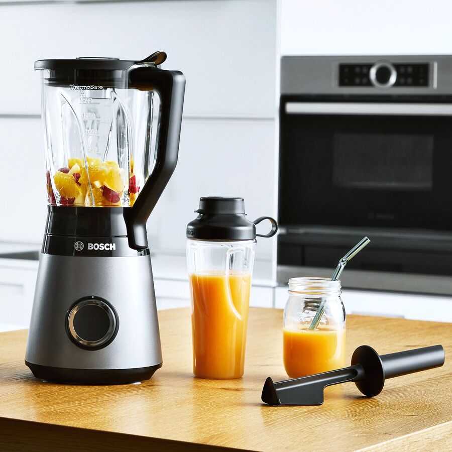 Bordblender med frugt i glaskanden på en træbordplade, med fyldt to-go-flaske ved siden af