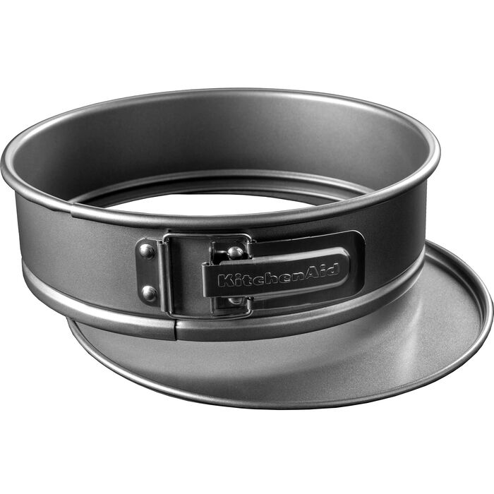 Rund springform i mørkegråt stål set skråt fra siden, med spændeklemme og KitchenAid-navnet præget på ringen og den løse bundplade stikkende lidt ud forneden, på hvid bund