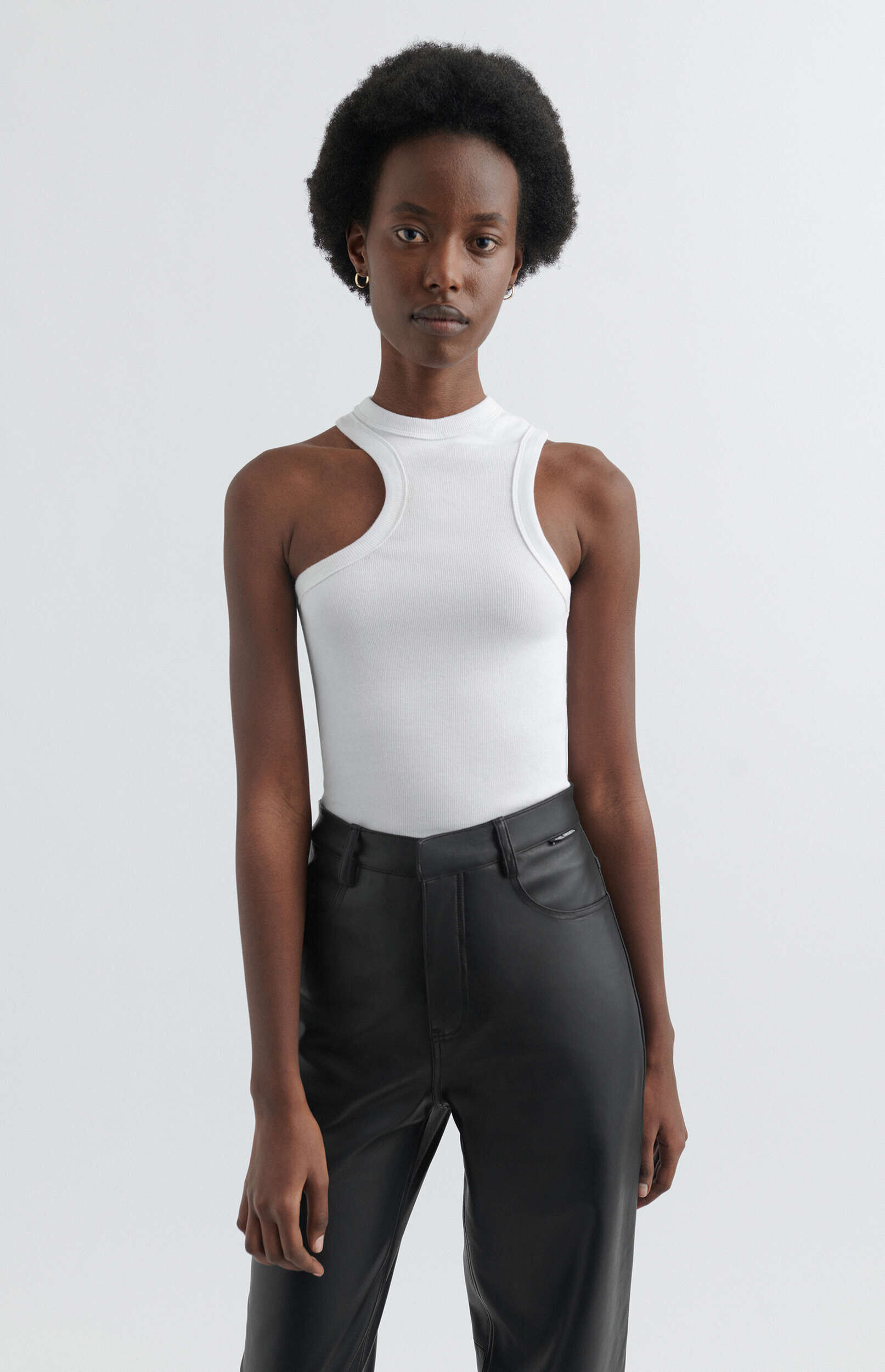 A young Black woman with short, curly dark hair stands against a plain white background, looking directly at the camera. She is wearing a fitted white ribbed tank top with an asymmetrical cutout over her right shoulder and black faux leather pants.