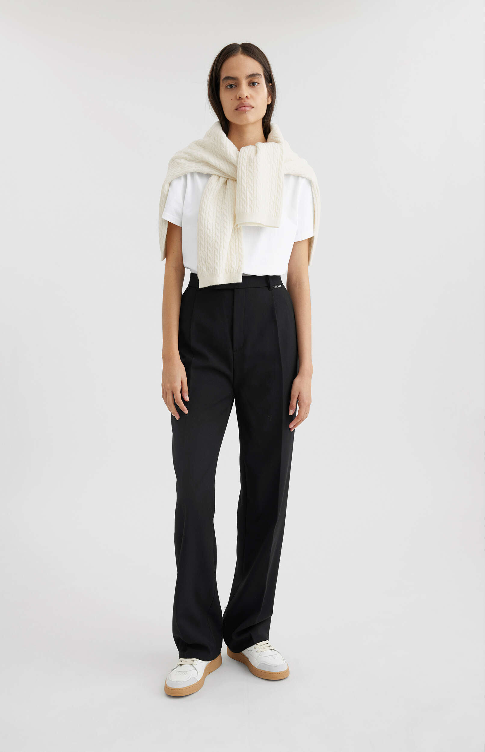 A full-length shot of a woman standing against a white background, wearing a white short-sleeved t-shirt, high-waisted black wide-leg trousers, and white sneakers with tan soles. A cream-colored cable-knit sweater is draped over her shoulders and tied in front. She has dark hair, is looking directly at the camera, and is in a relaxed pose.