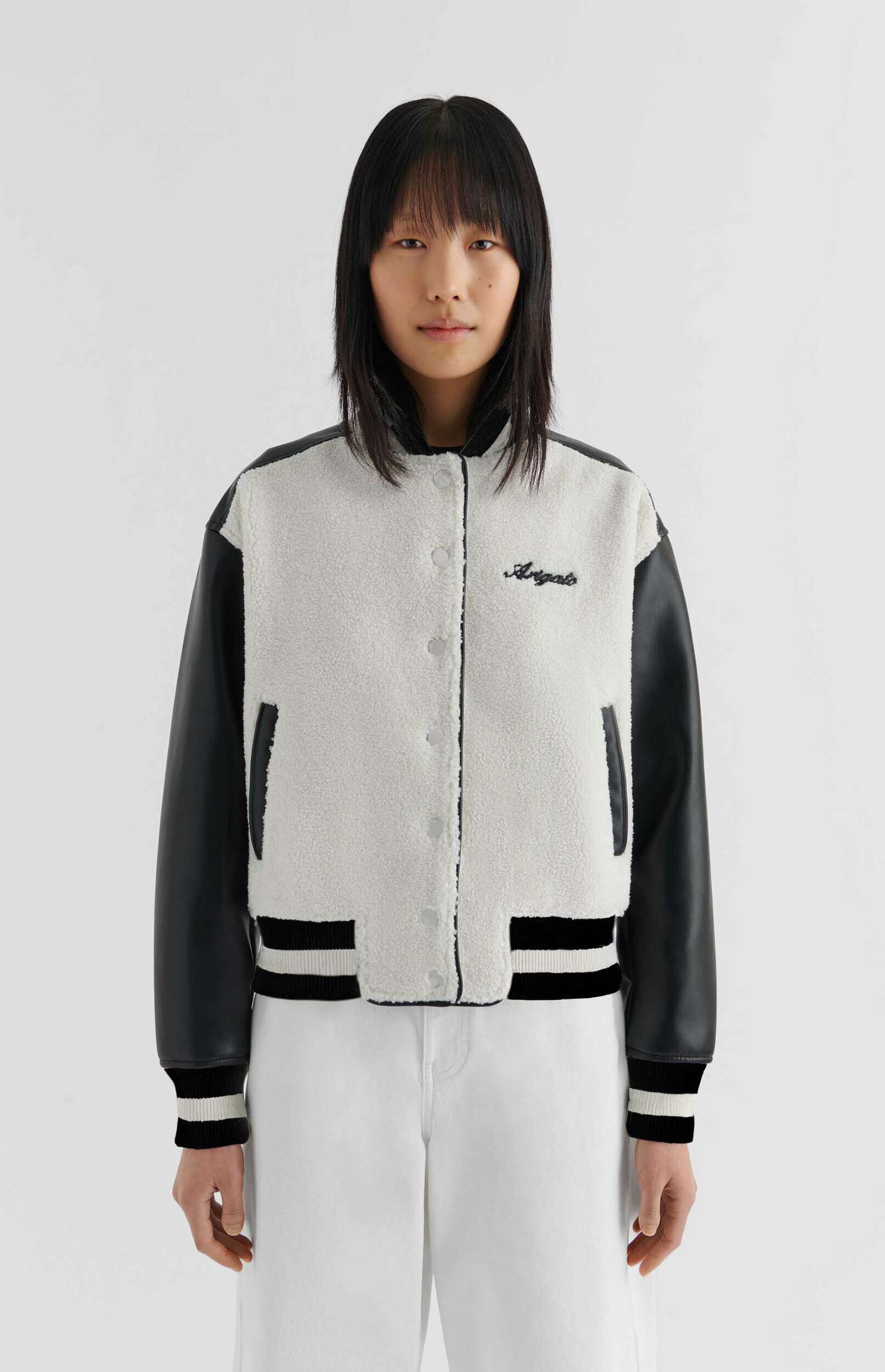 A young person with straight dark hair and bangs stands against a white background, wearing an off-white sherpa and black leather-sleeved bomber jacket. The jacket features black and white striped ribbed cuffs and hem, and black embroidery that reads 'Arigato' on the chest. They are also wearing white pants.