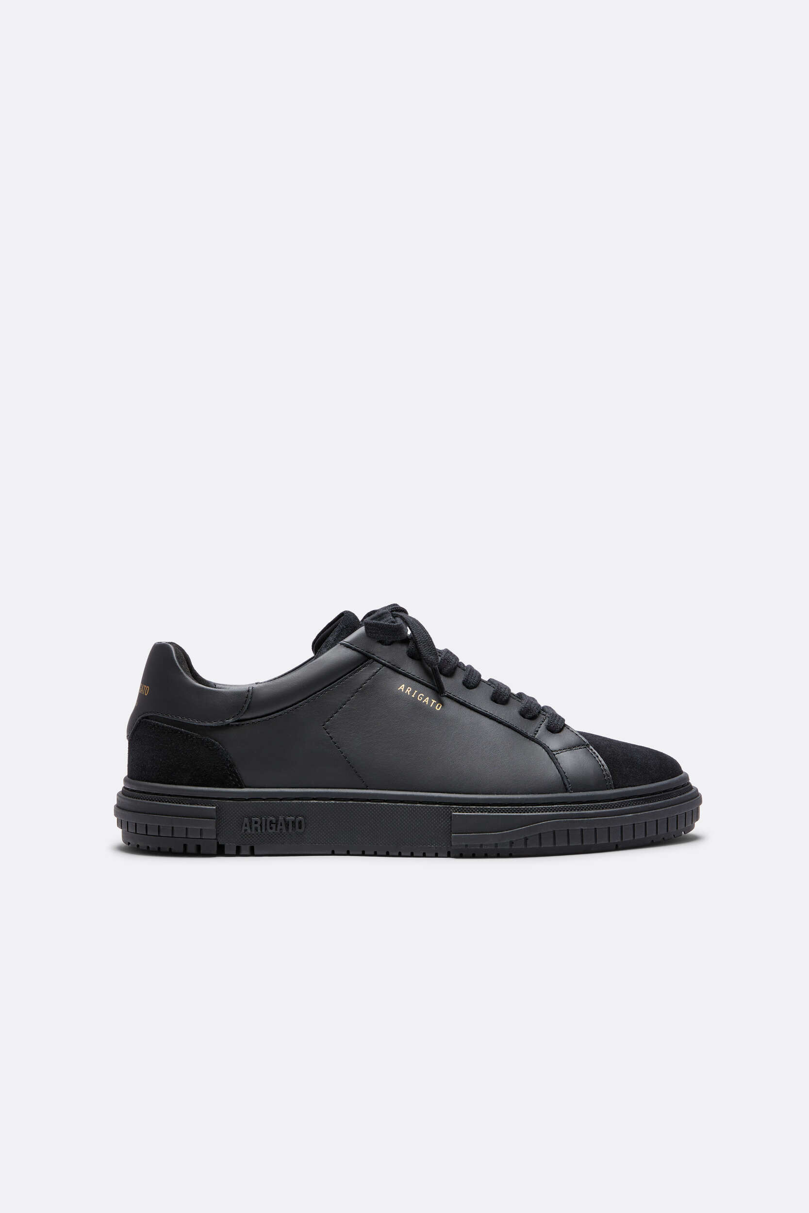 A single black low-top sneaker from Axel Arigato, featuring smooth leather and suede panels. The 'ARIGATO' logo is visible in gold on the side and embossed on the black rubber sole. The shoe has black laces and a chunky black sole with textured details, set against a plain light grey background.
