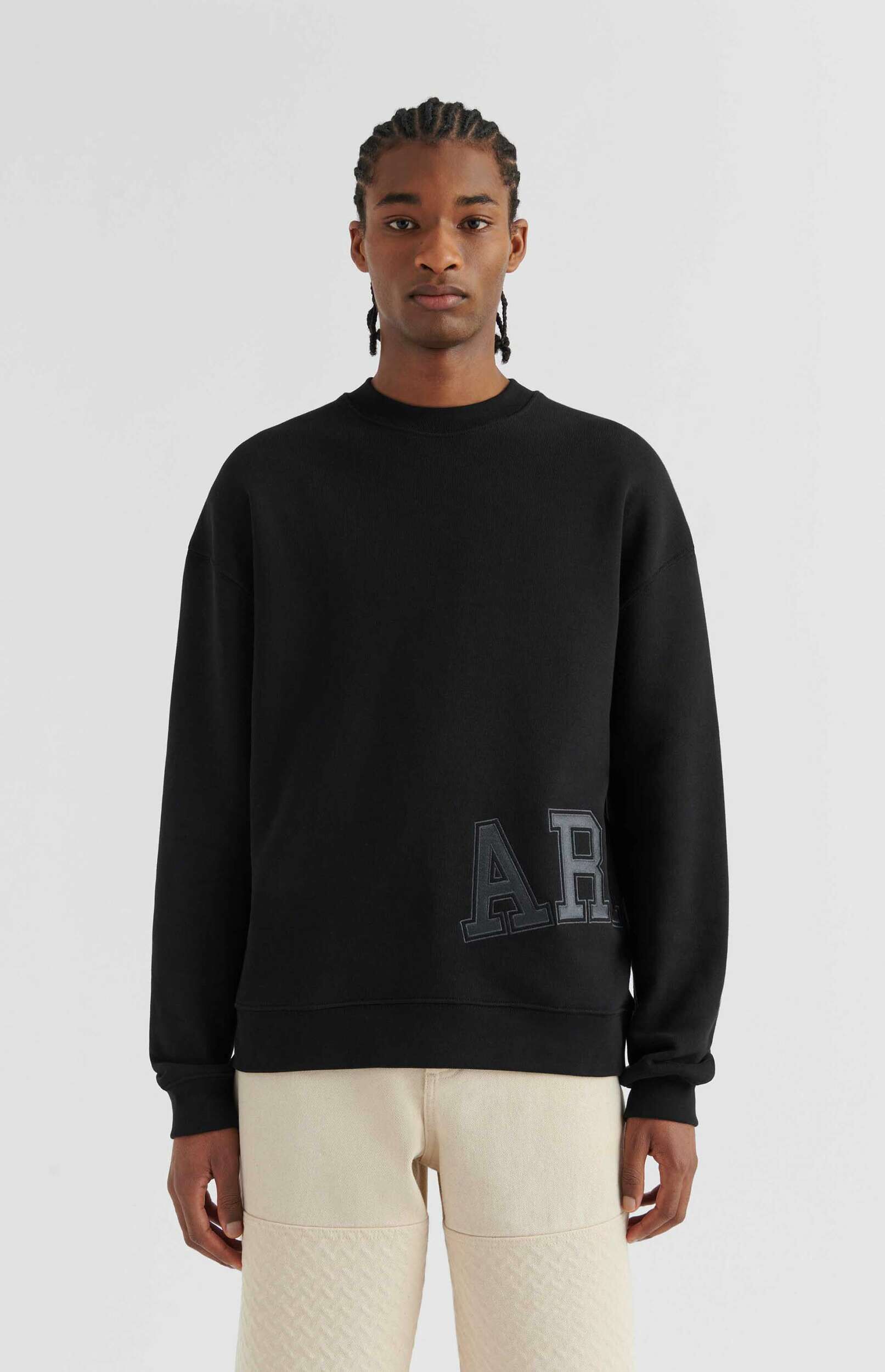 A man wearing a black crewneck sweatshirt with 'AR' letters and light-colored pants stands against a white background.