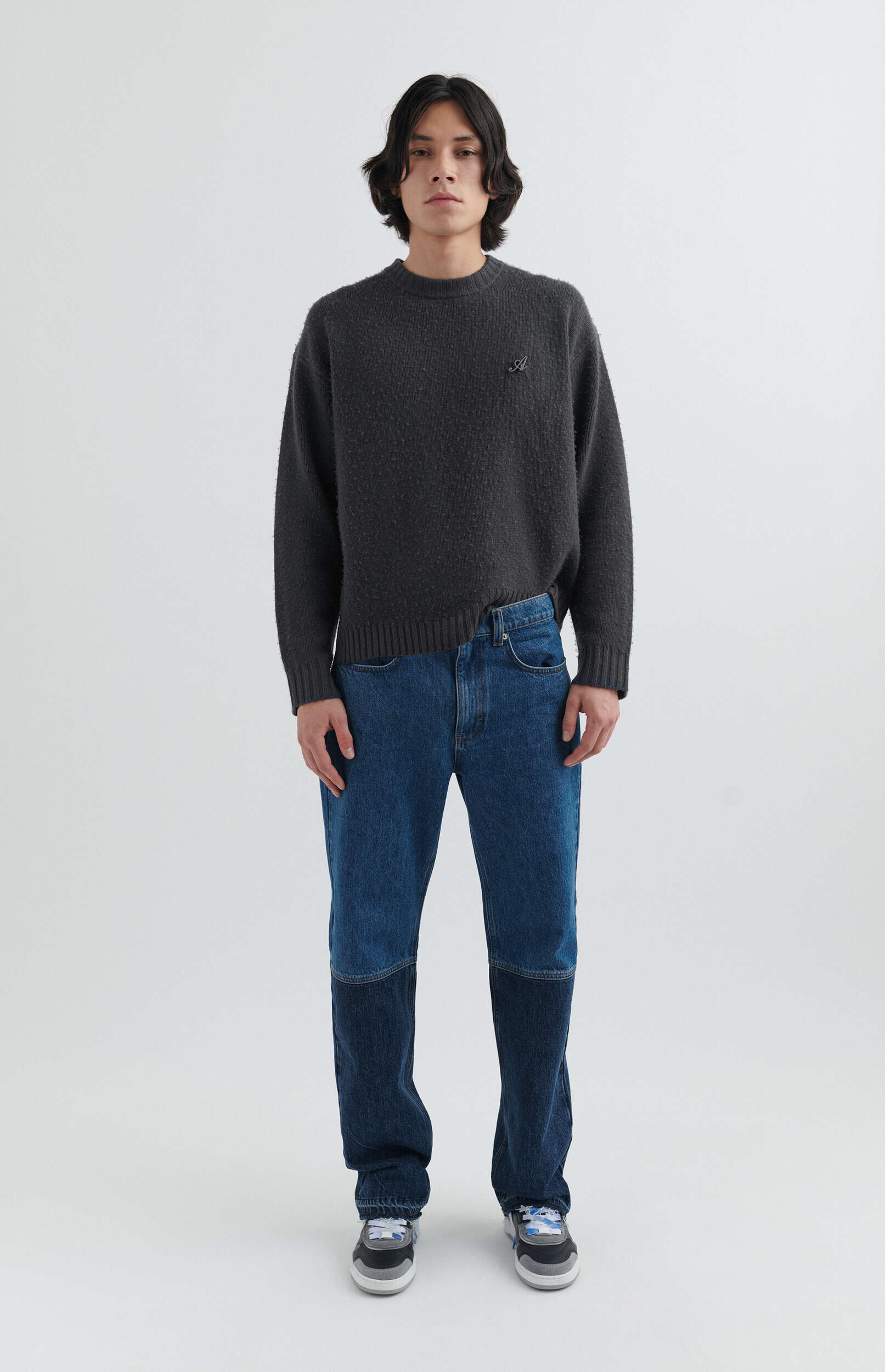 A full-body shot of a person with dark, wavy hair standing against a plain white background. They are wearing a dark charcoal gray knitted sweater with a small embroidered 'A' on the left chest, two-tone blue jeans with darker patches on the lower legs, and multi-colored sneakers with blue laces.