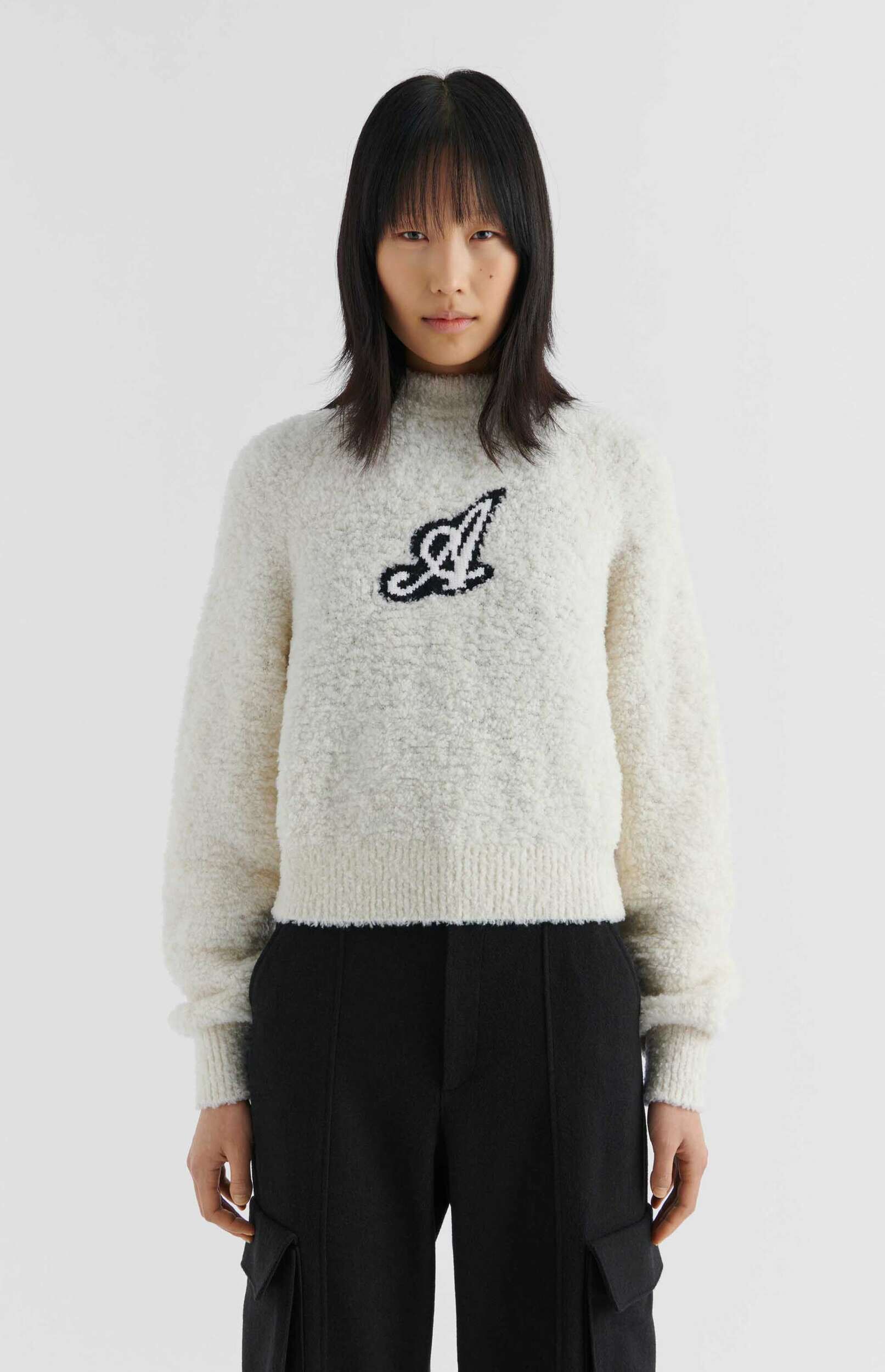 A woman with dark hair and bangs wears a cream-colored, textured mock-neck sweater featuring a black and white stylized 'A' logo on the chest, paired with black wide-leg cargo pants. She stands facing forward against a plain white background.