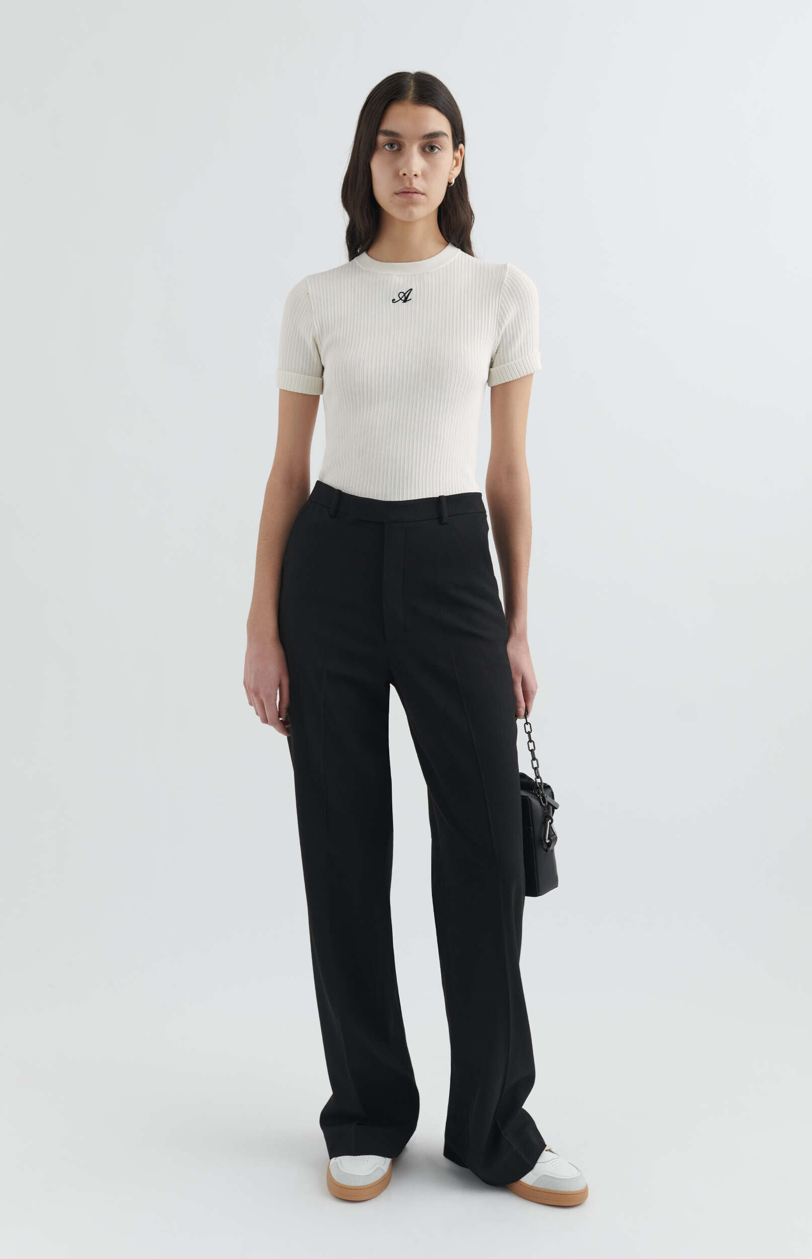 A woman stands in a studio, wearing a cream ribbed short-sleeve top with a black 'A' logo, black wide-leg trousers, and white sneakers with brown soles. She holds a small black shoulder bag with a chain strap.