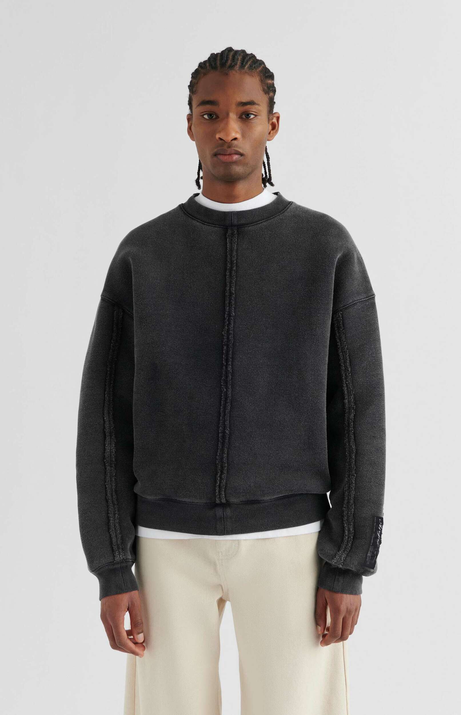 A man wearing a dark grey, distressed-look sweatshirt with prominent exposed seams down the front and along the arms, layered over a white t-shirt and paired with light beige wide-leg trousers. He has braided hair and is standing against a plain white background.