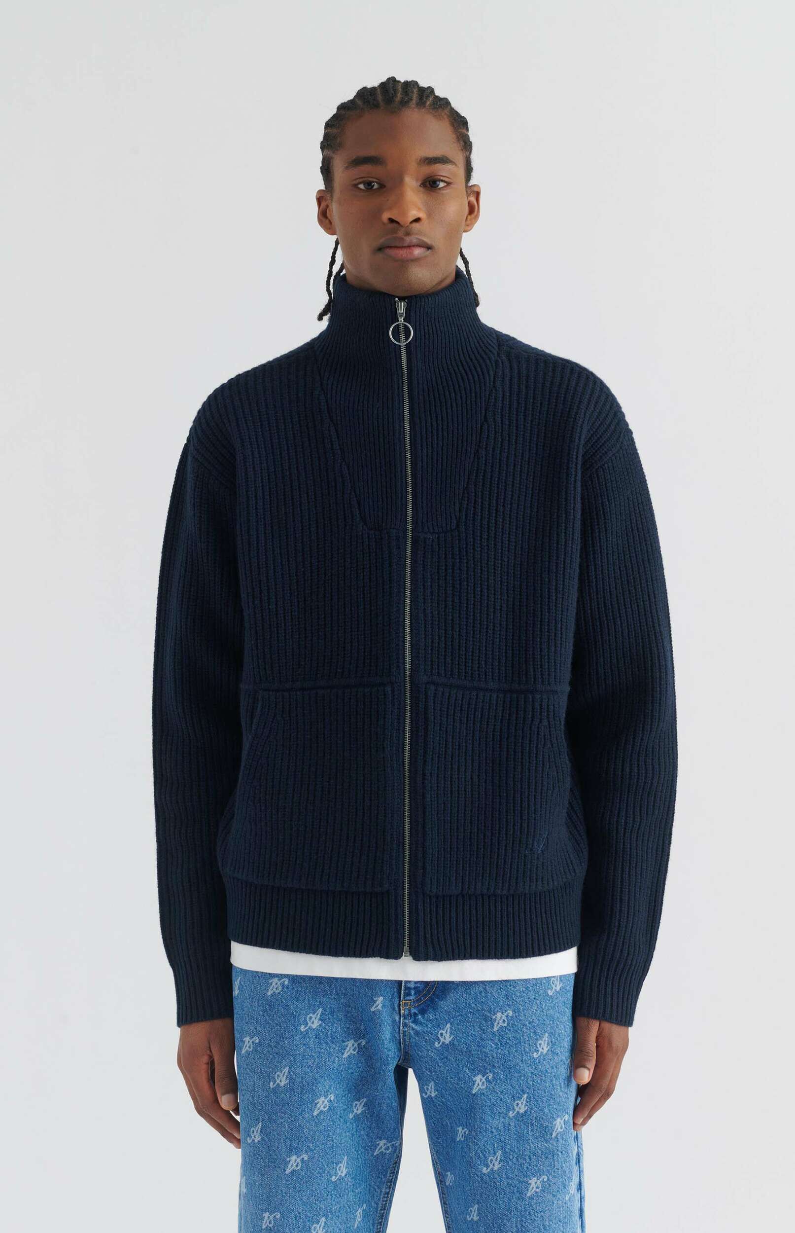 A man with braided hair stands against a white background, wearing a dark navy blue ribbed knit zipped cardigan with a high collar and two front pockets, a white t-shirt, and light blue denim jeans with a repeating white 'A' pattern.