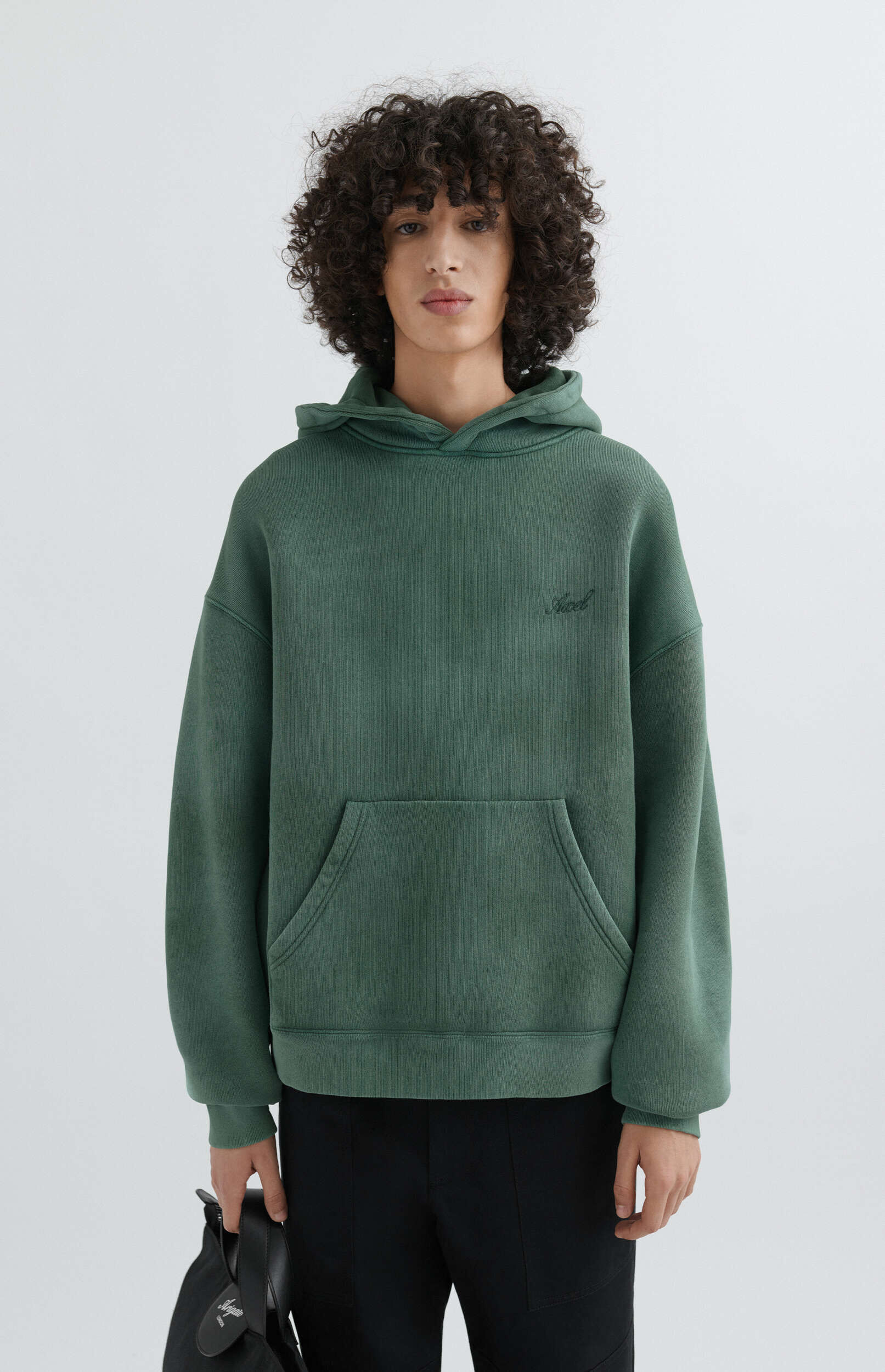 A young man with curly hair wearing a dark green oversized hoodie with a small embroidered logo, black pants, and holding a black bag in his left hand, standing against a white background.