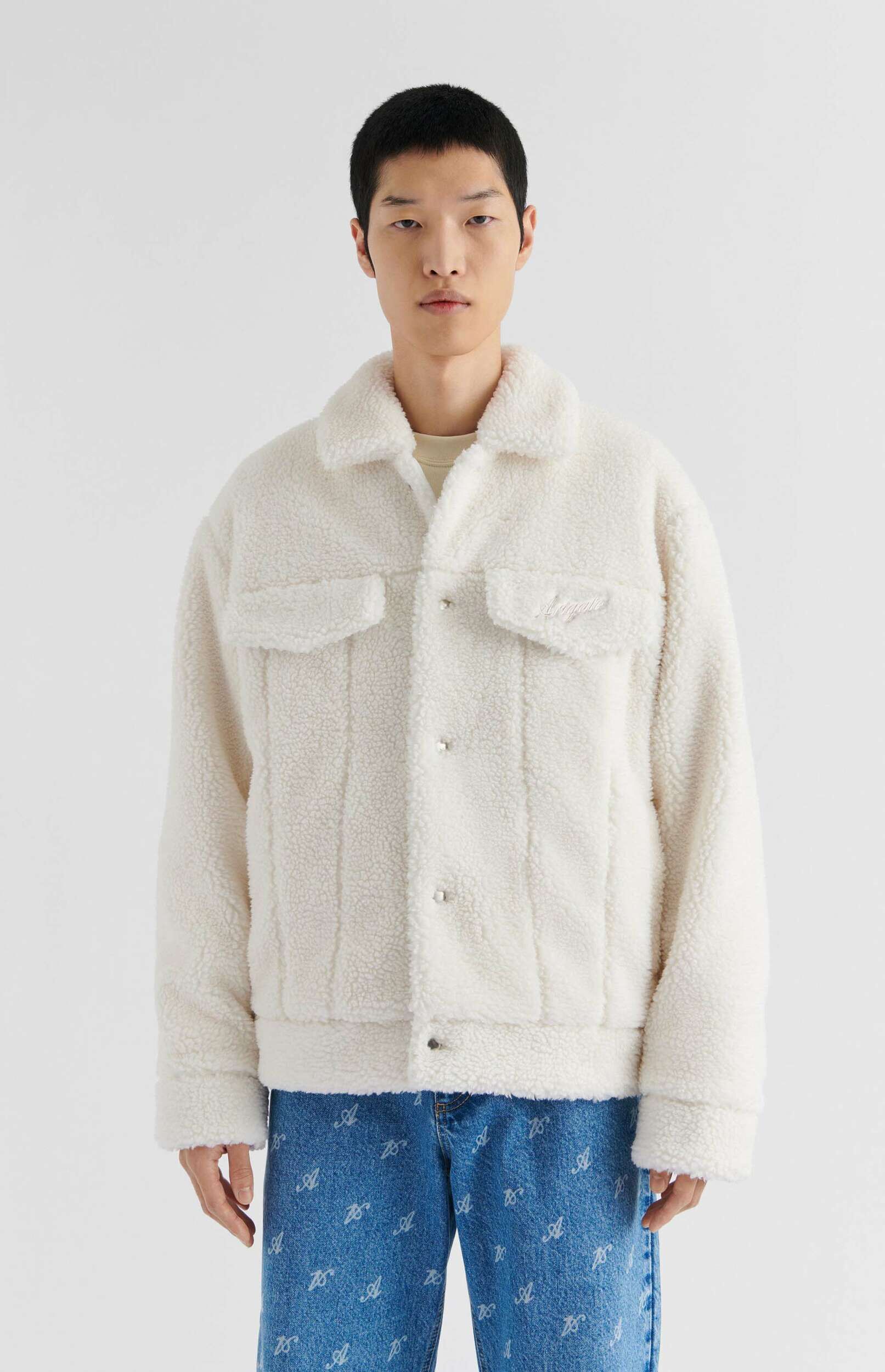 A male model stands against a plain white background, wearing a creamy white sherpa fleece jacket with a collar, two chest pockets (one with a subtle embroidered logo), and silver snap buttons. He pairs it with blue denim jeans featuring a repeating pattern of white 'A' letters. The model has short dark hair and a neutral expression.