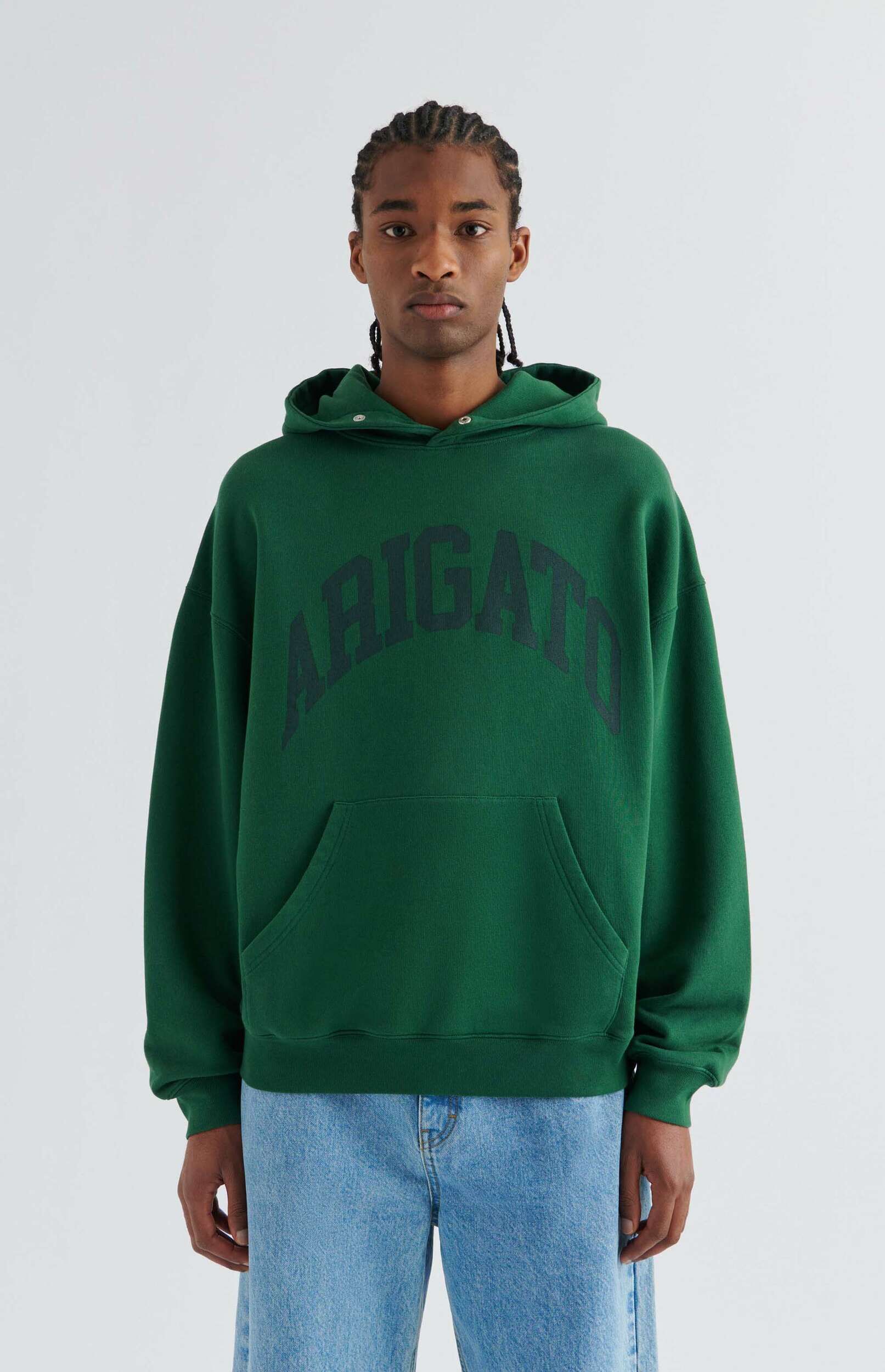 A person with dreadlocks wearing a forest green oversized hoodie with "ARIGATO" in dark letters across the chest, paired with light wash blue jeans, standing against a white background.