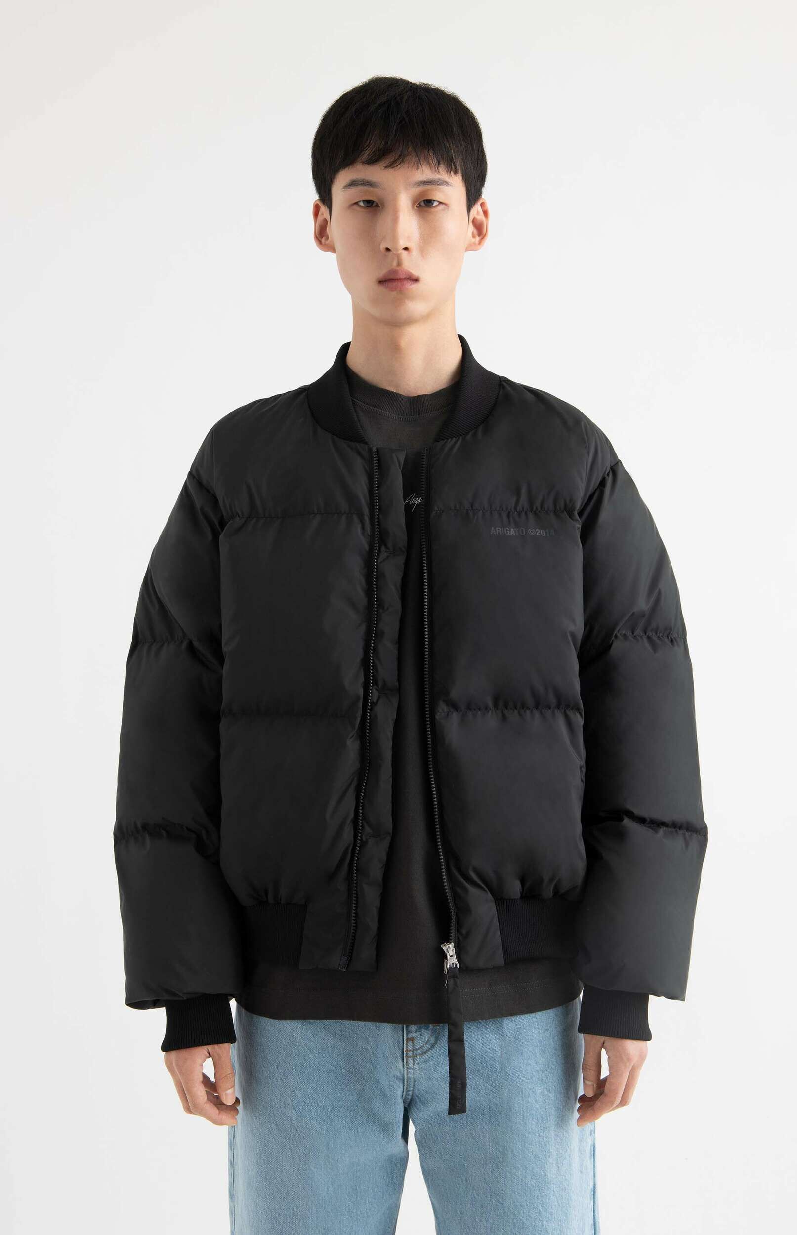 A young man wearing a black puffer bomber jacket with 'ARIGATO ©2014' text, a dark t-shirt, and light blue jeans, standing against a white background.