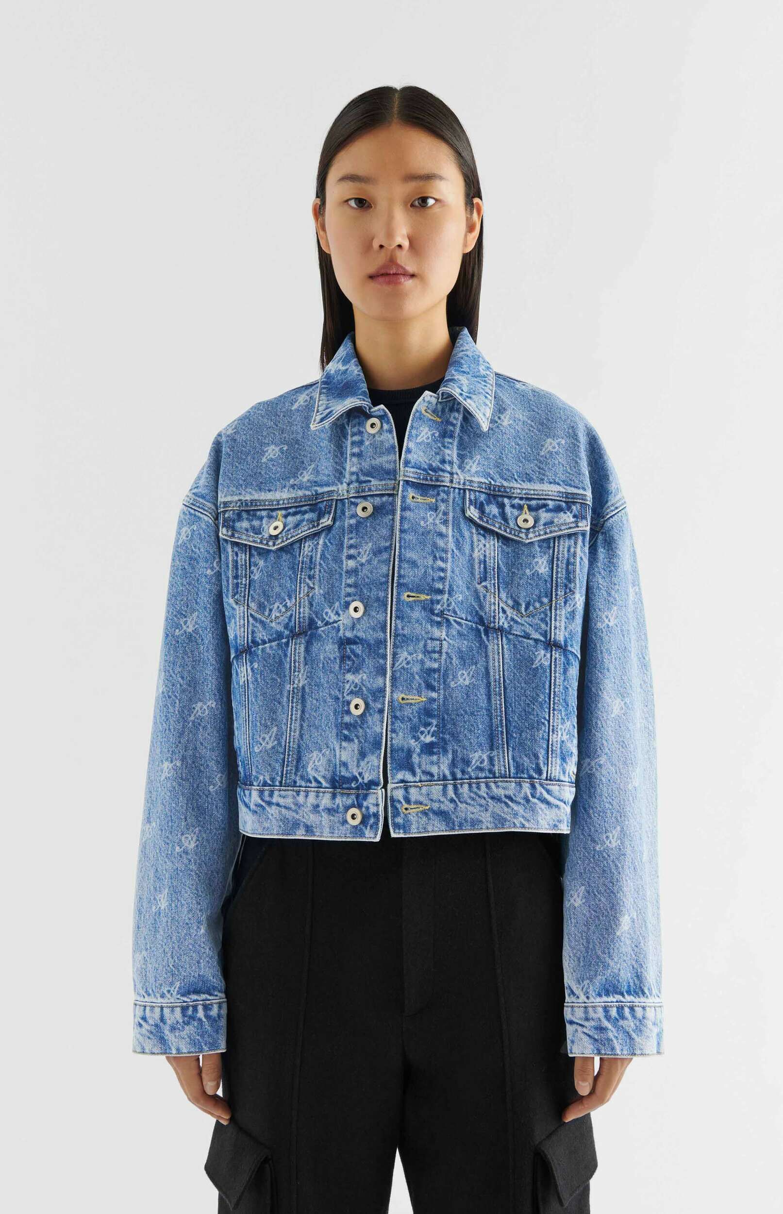 A person with long dark hair stands against a white background, wearing a light blue, acid-wash denim jacket with an all-over white abstract pattern, a black crew neck top, and black wide-leg trousers.