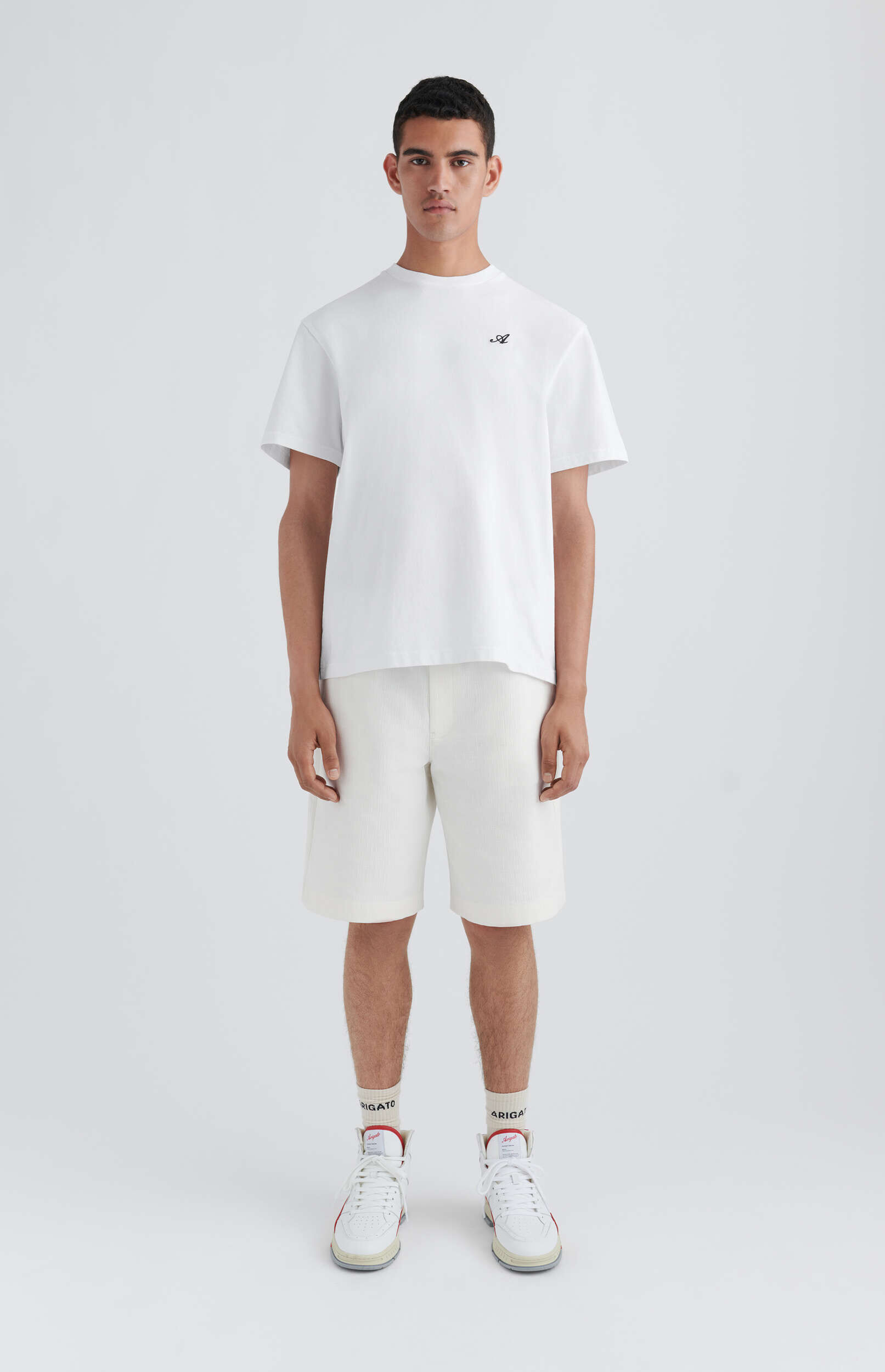 A young man stands full-body facing forward, wearing a plain white short-sleeved t-shirt with a small dark script logo on the left chest, light cream-colored textured shorts, white high-top sneakers with red accents, and white crew socks with black 'RIGATO' text. The background is a clean white studio setting.