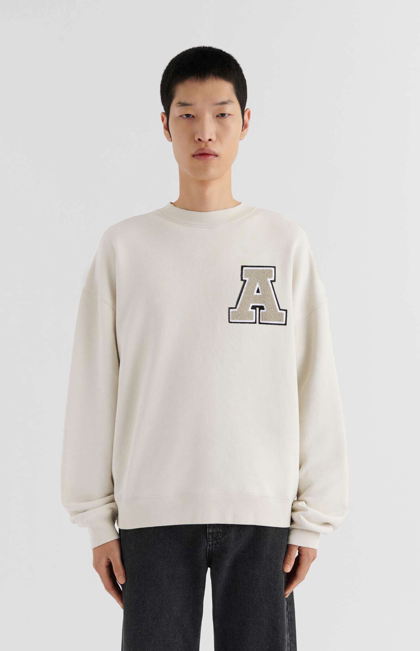 A young East Asian man stands facing forward against a plain white background, wearing a light cream or off-white oversized crewneck sweatshirt. The sweatshirt features a large, textured patch of the capital letter 'A' in a beige color with a dark outline on the left chest. He is also wearing dark grey or black wide-leg trousers. The man has short, dark hair and a neutral expression.