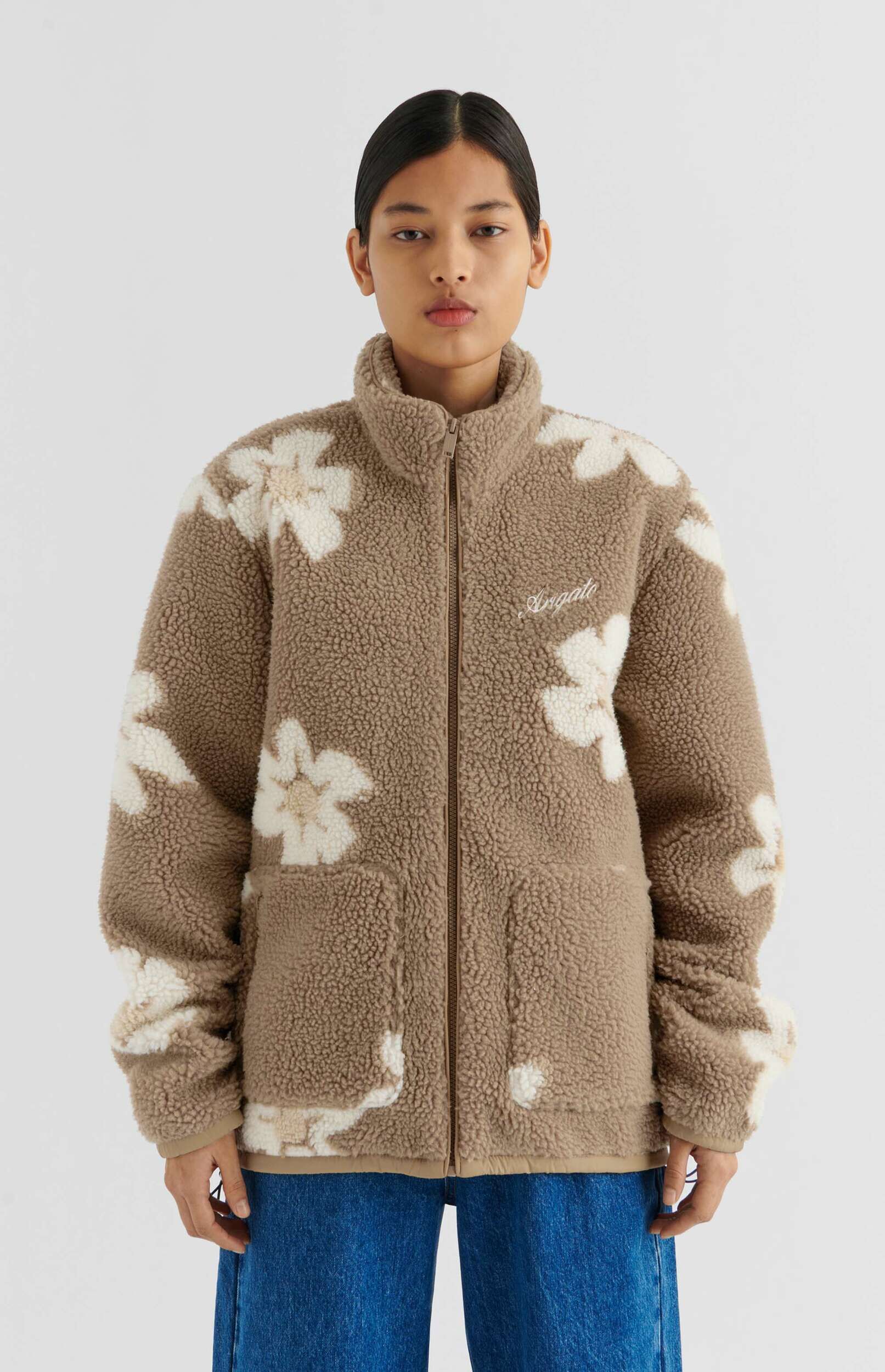 A person with dark hair pulled back, wearing a cozy brown sherpa fleece jacket with large white floral patterns, and blue jeans, standing against a plain white background.