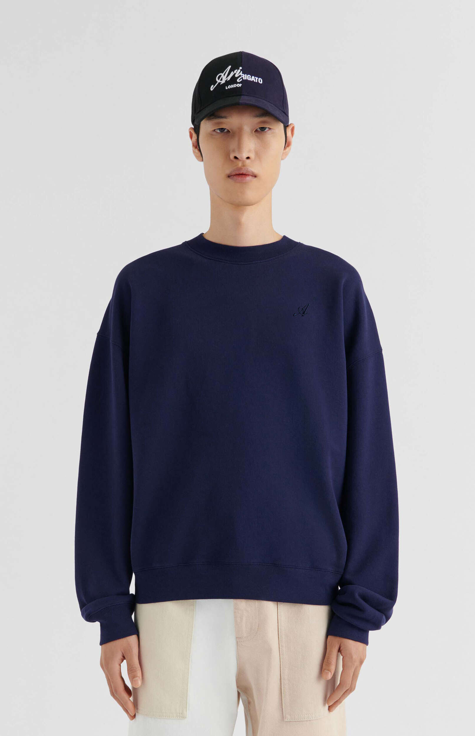 A male model stands against a white background, wearing a navy blue crew-neck sweatshirt with a subtle 'A' logo, light beige and white color-block pants, and a dark baseball cap with 'ARIGATO LONDON' in white script.