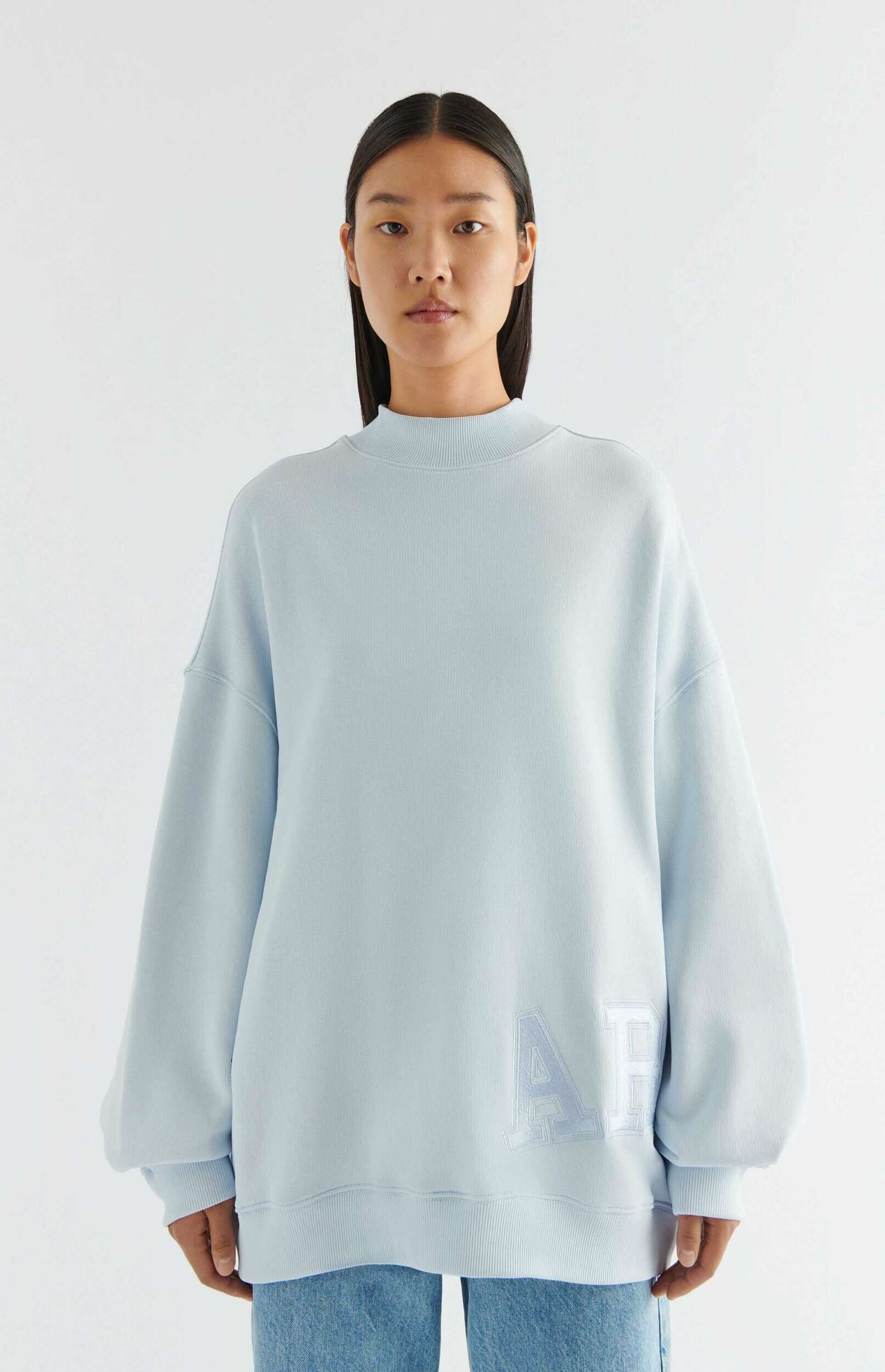 A woman wearing a light blue oversized sweatshirt with a mock neck and ribbed cuffs, featuring a subtle embroidered "AP" logo on the lower right. She has long dark hair and is wearing blue jeans, set against a white background.