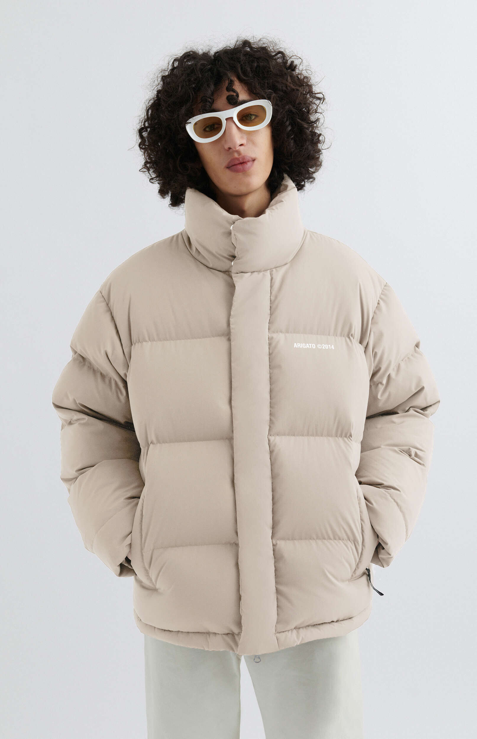 A person with curly dark hair wearing a beige puffer jacket and white-framed sunglasses with yellow lenses, hands in pockets, standing against a light background.