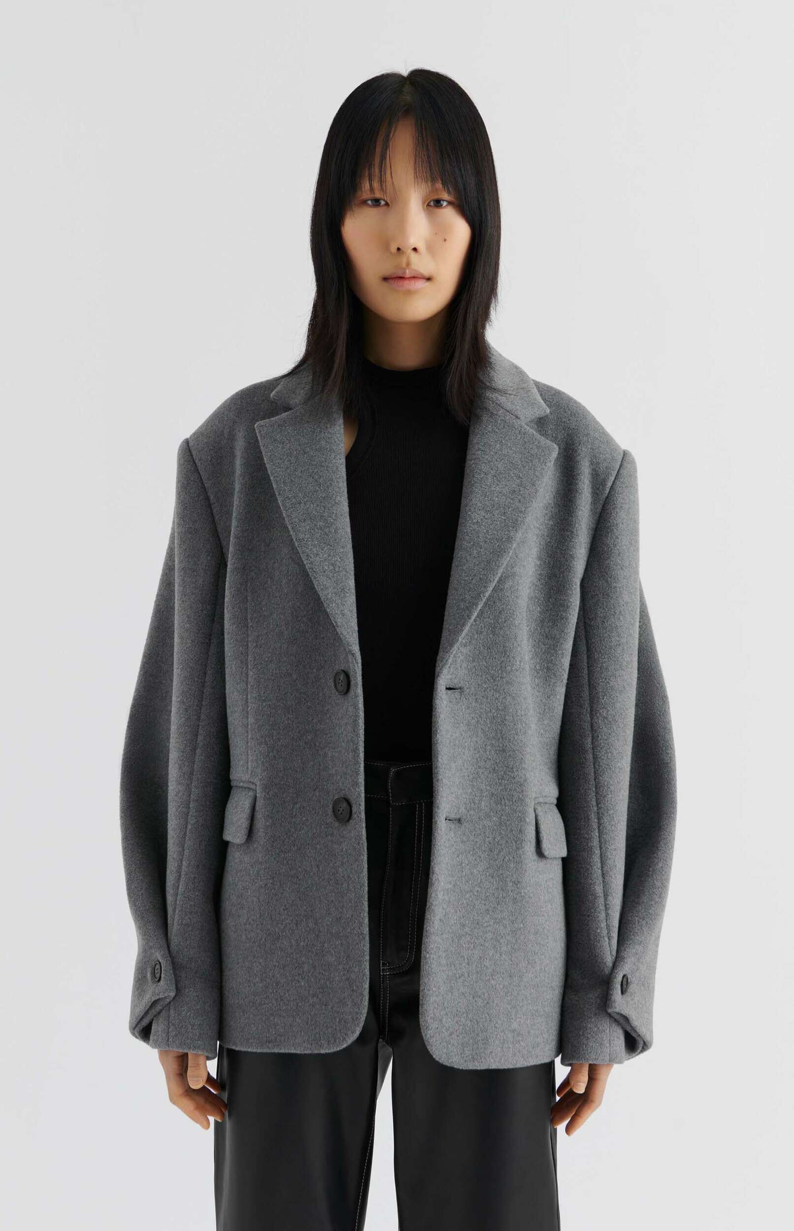 A person with black hair and bangs poses against a white background, wearing a charcoal grey oversized blazer over a black ribbed high-neck top and black leather-look trousers.