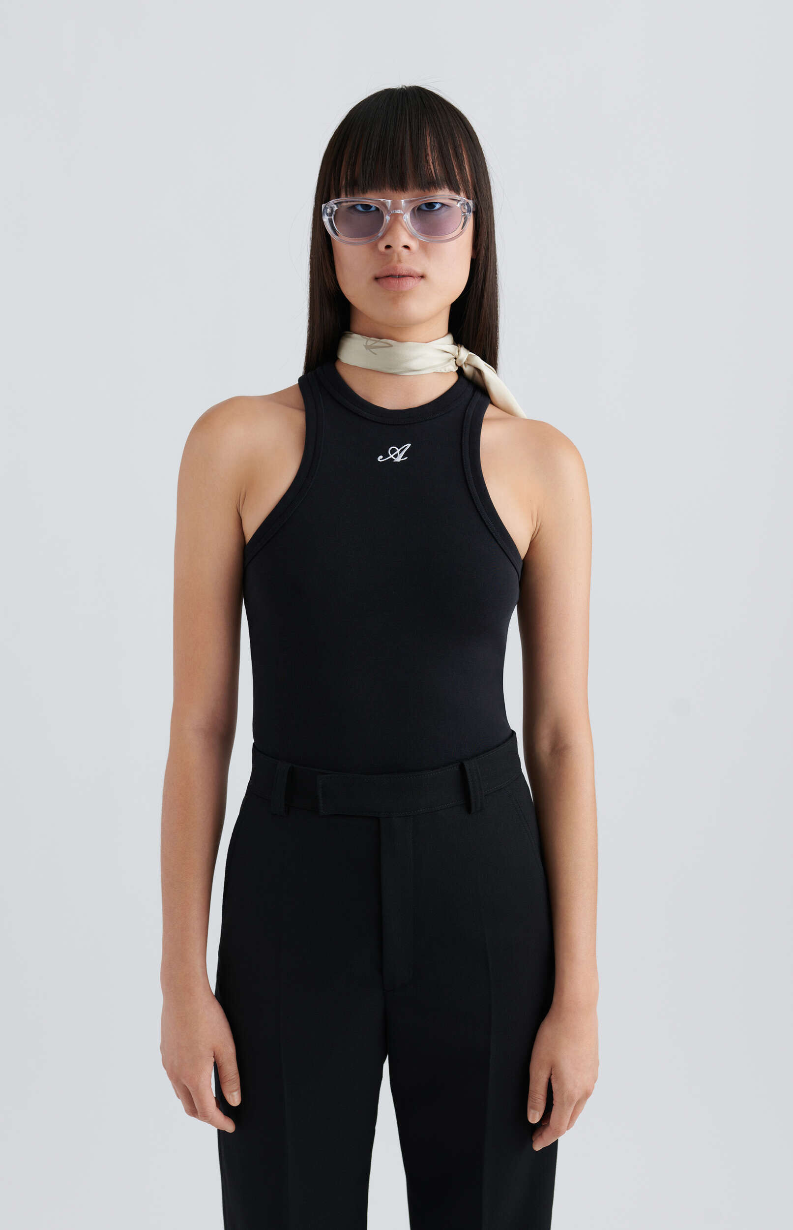 A person with long dark hair and bangs, wearing clear-framed sunglasses with purple tinted lenses, a black ribbed tank top with a white script logo, black trousers, and a light beige silk scarf around their neck, stands against a plain light gray background.