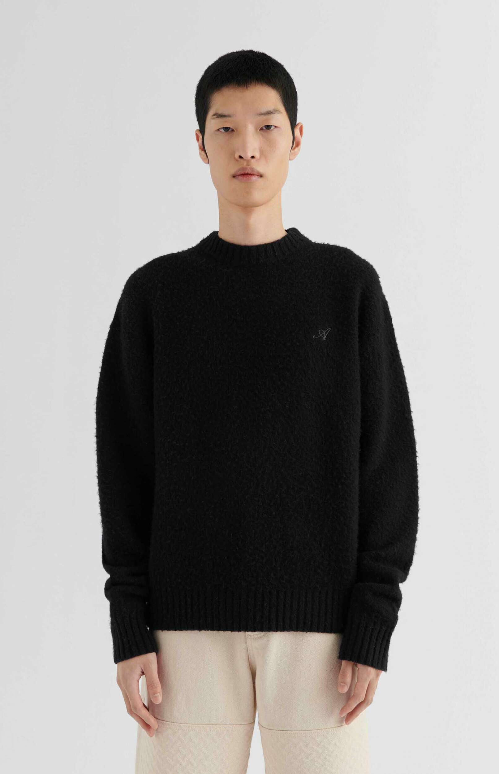 A person wearing a black textured crewneck sweater with a small, stylized gray 'A' logo embroidered on the left chest, paired with light beige pants. The person has short dark hair and stands against a plain white background.