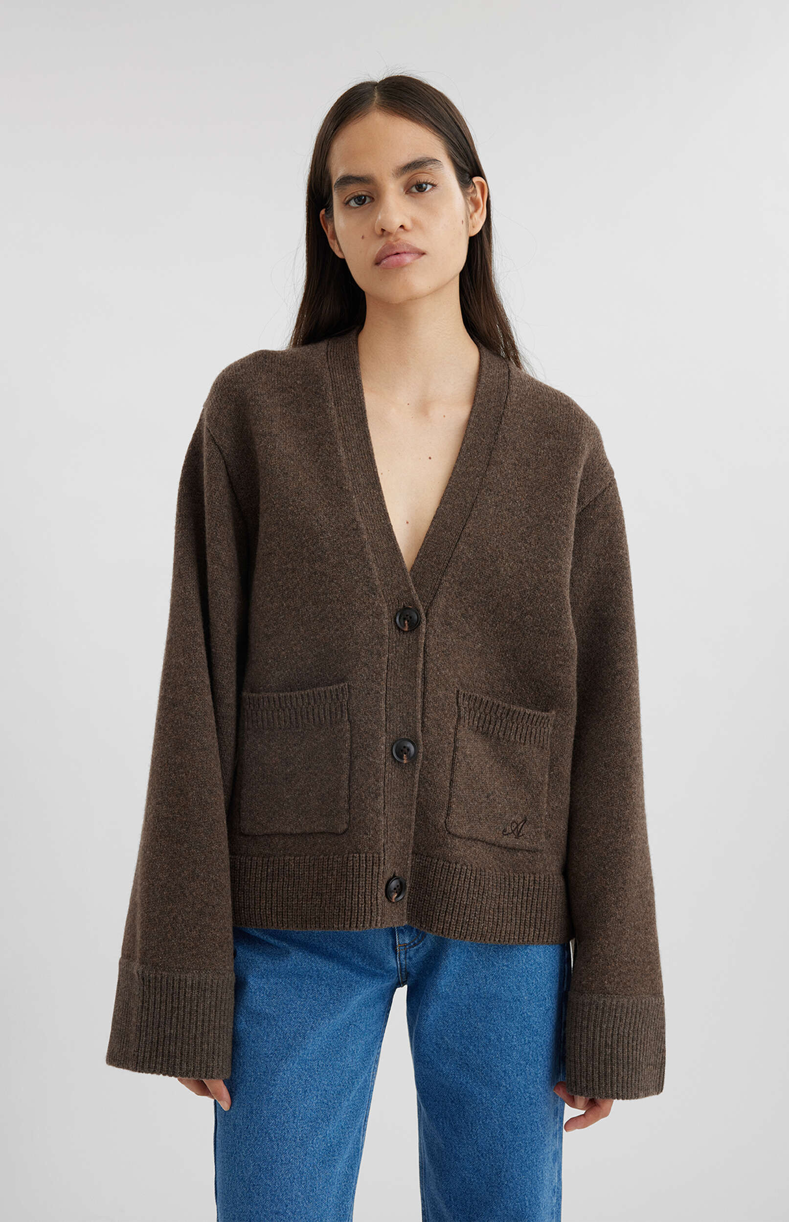A woman with long dark hair and a natural look stands against a white background, wearing a loose-fitting, V-neck brown knitted cardigan with three dark buttons and two large front pockets. She is also wearing blue denim jeans. The cardigan features ribbed cuffs and hem, and a subtle embroidered 'A' on the right pocket.