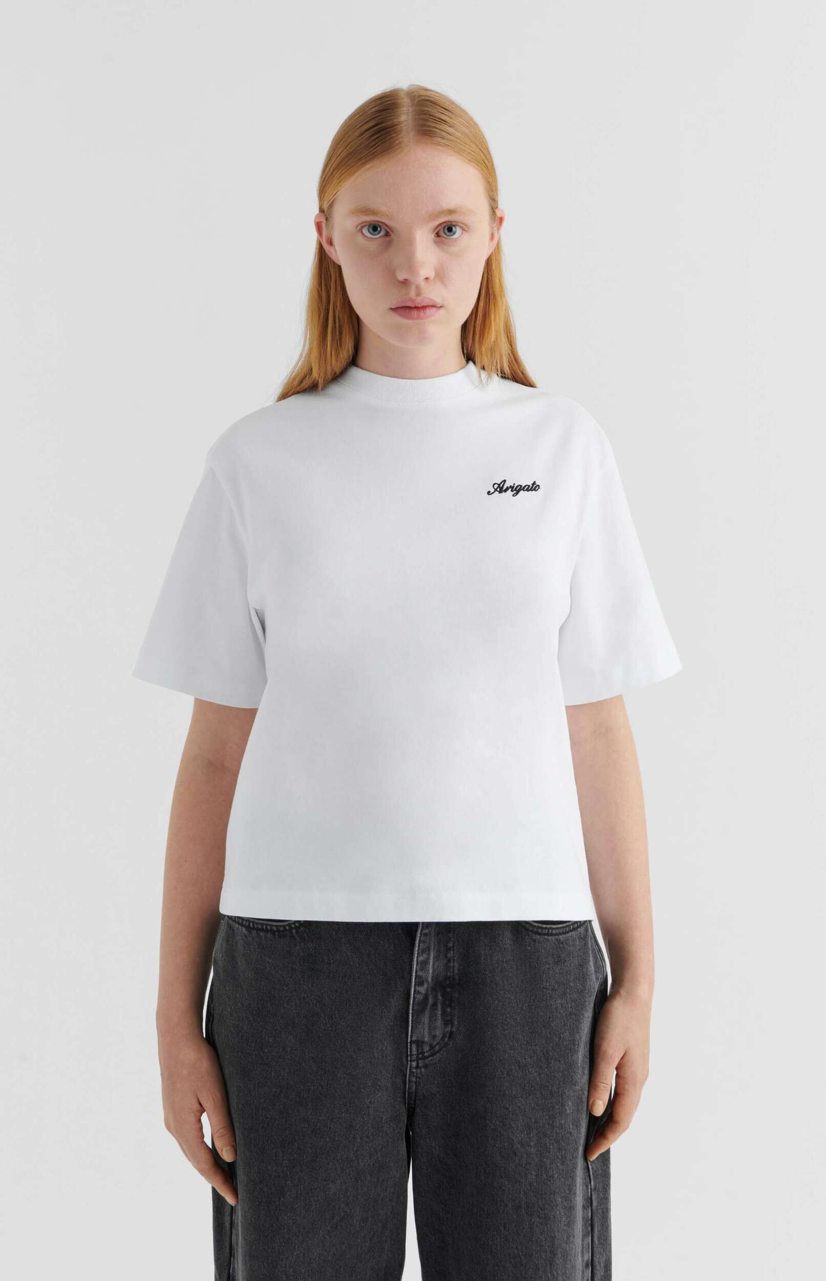 A young woman with long, light reddish-blonde hair and blue eyes stands facing forward, wearing a white crew-neck t-shirt with 'Arigato' in black script on the chest and dark gray jeans, against a plain white background.