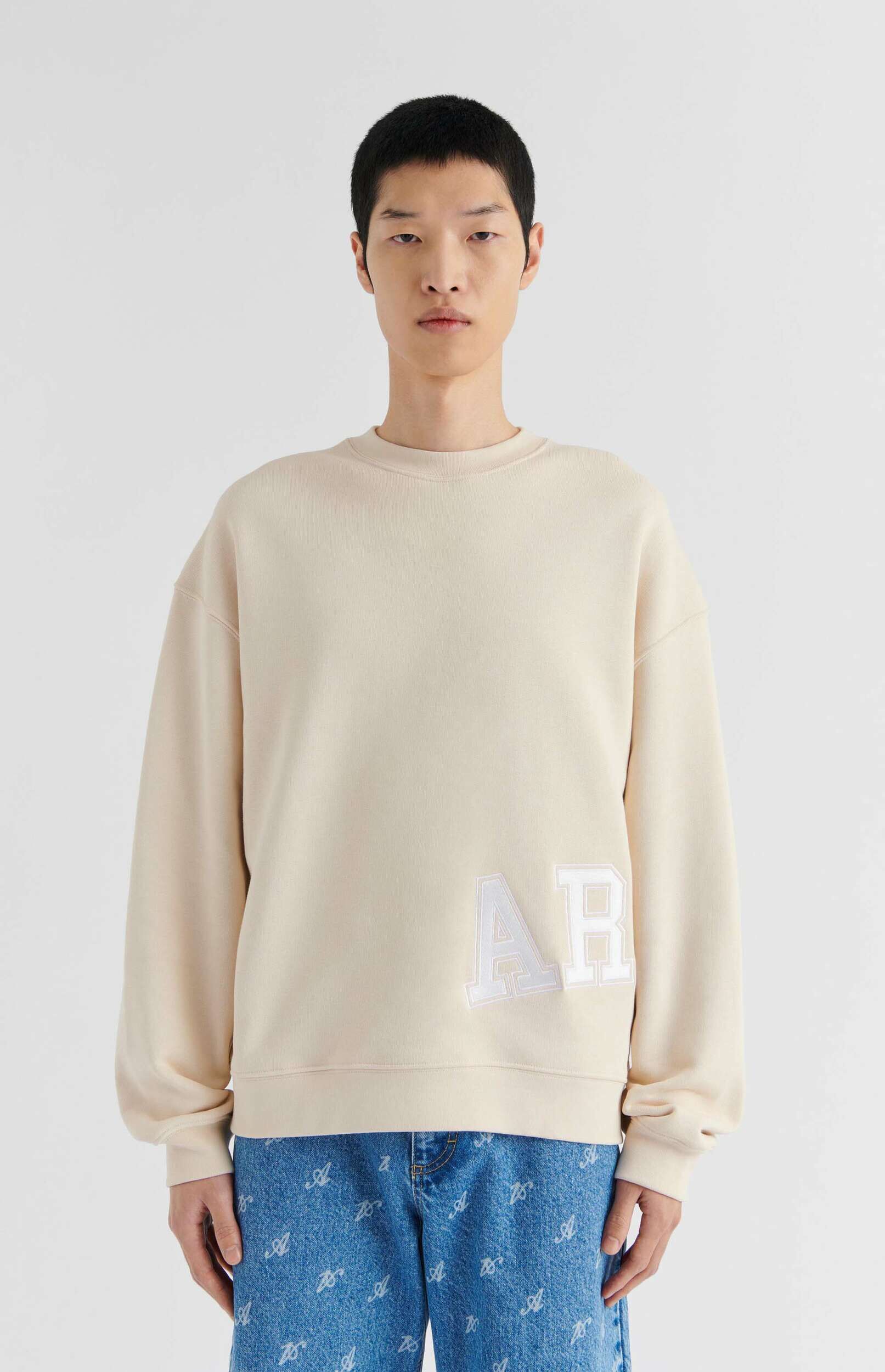 A person wearing a light beige oversized sweatshirt with large white 'AR' letters on the front, paired with blue denim jeans featuring a repeating pattern of white script 'A's. The person is standing front-facing against a plain white background.