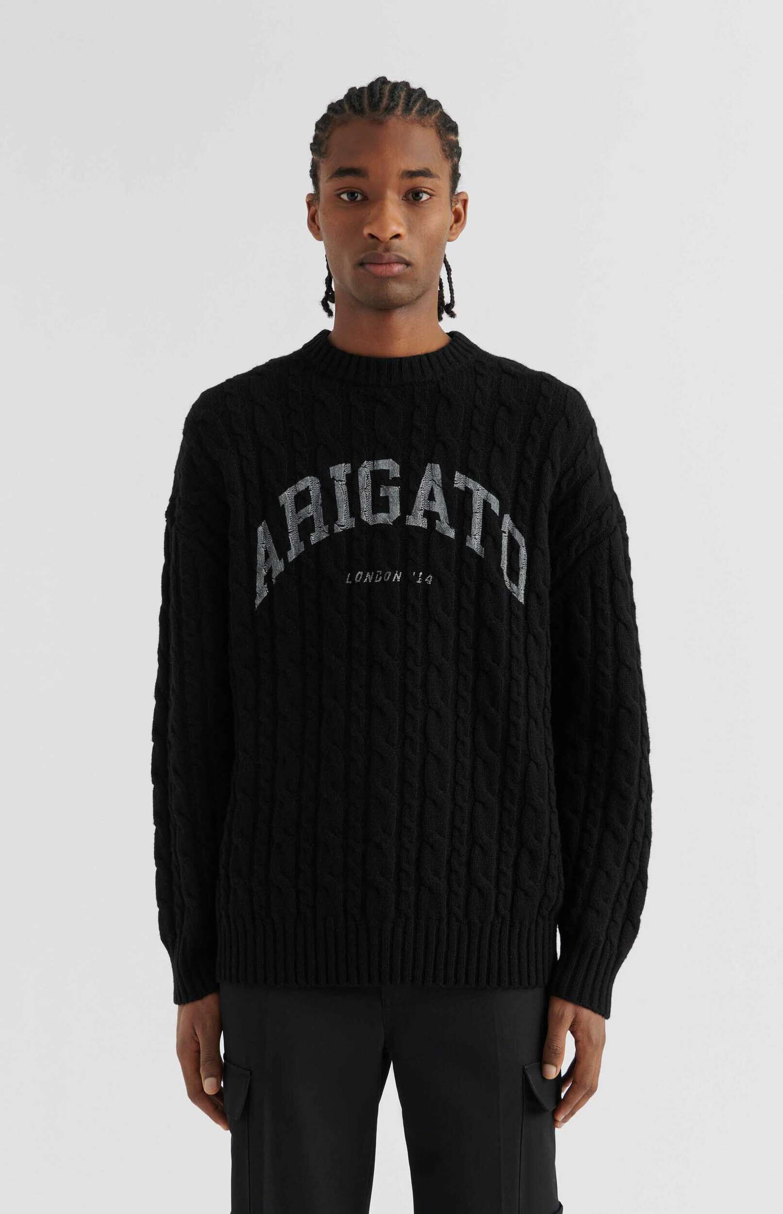 A man with braids wears a black cable-knit sweater featuring 'ARIGATO' in distressed silver lettering and 'LONDON '14' below it, paired with black pants, standing against a white background.