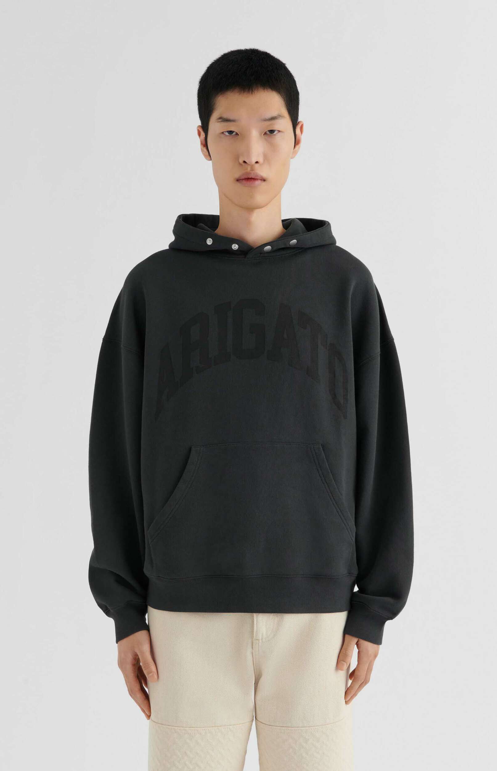 A young East Asian man with short dark hair stands facing forward, wearing a dark charcoal grey oversized hoodie with the word 'ARIGATO' embossed in matching dark letters across the chest. The hoodie features a kangaroo pocket and silver snap buttons along the hood. He is also wearing light beige trousers. The background is a plain white studio setting.