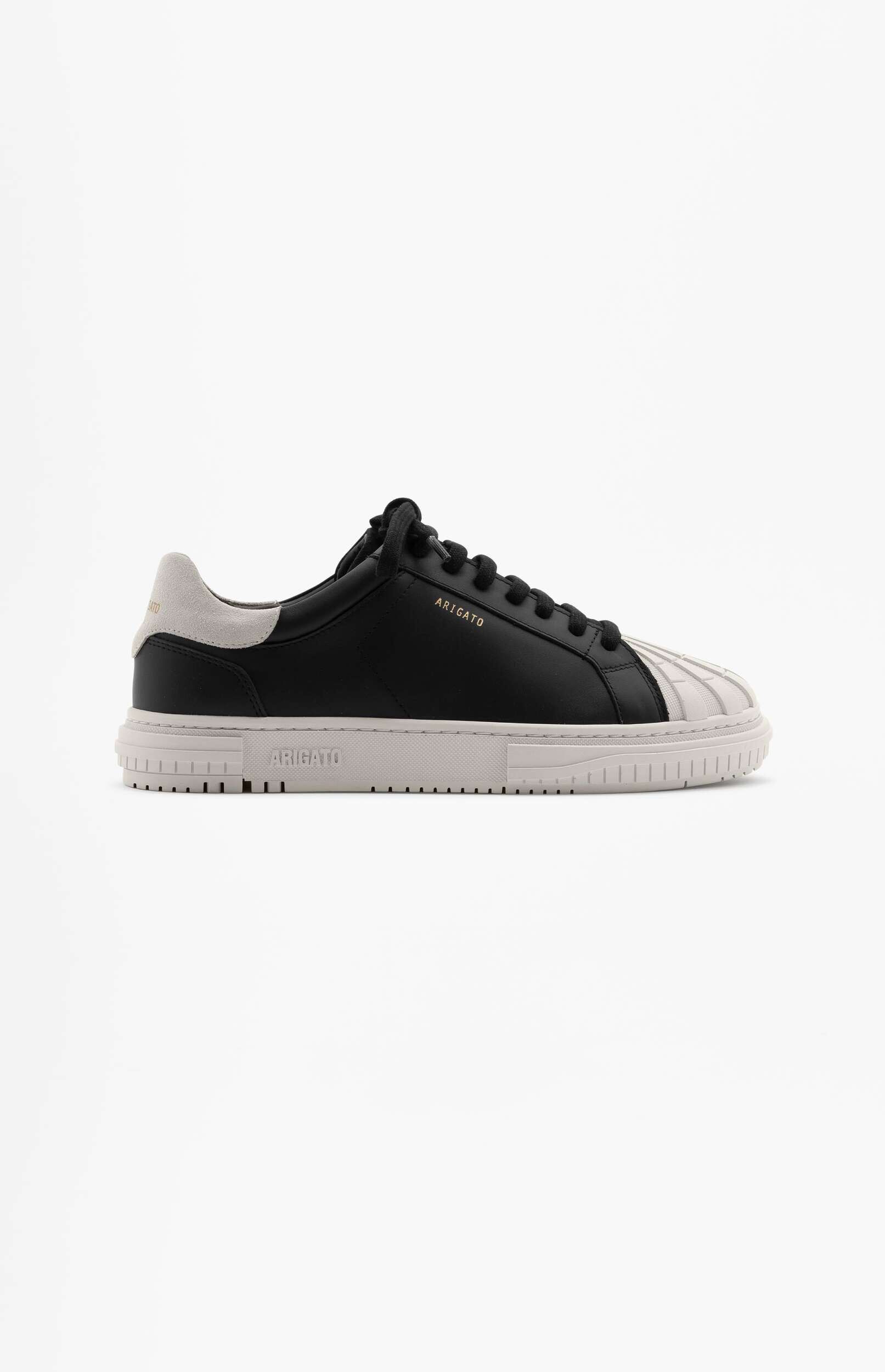 A single black and light grey Axel Arigato sneaker is displayed against a plain white background. The shoe features a black leather upper, black laces, a light grey heel counter, and a distinctive ribbed light grey toe cap. The brand name 'ARIGATO' is visible in gold lettering on the side of the shoe.