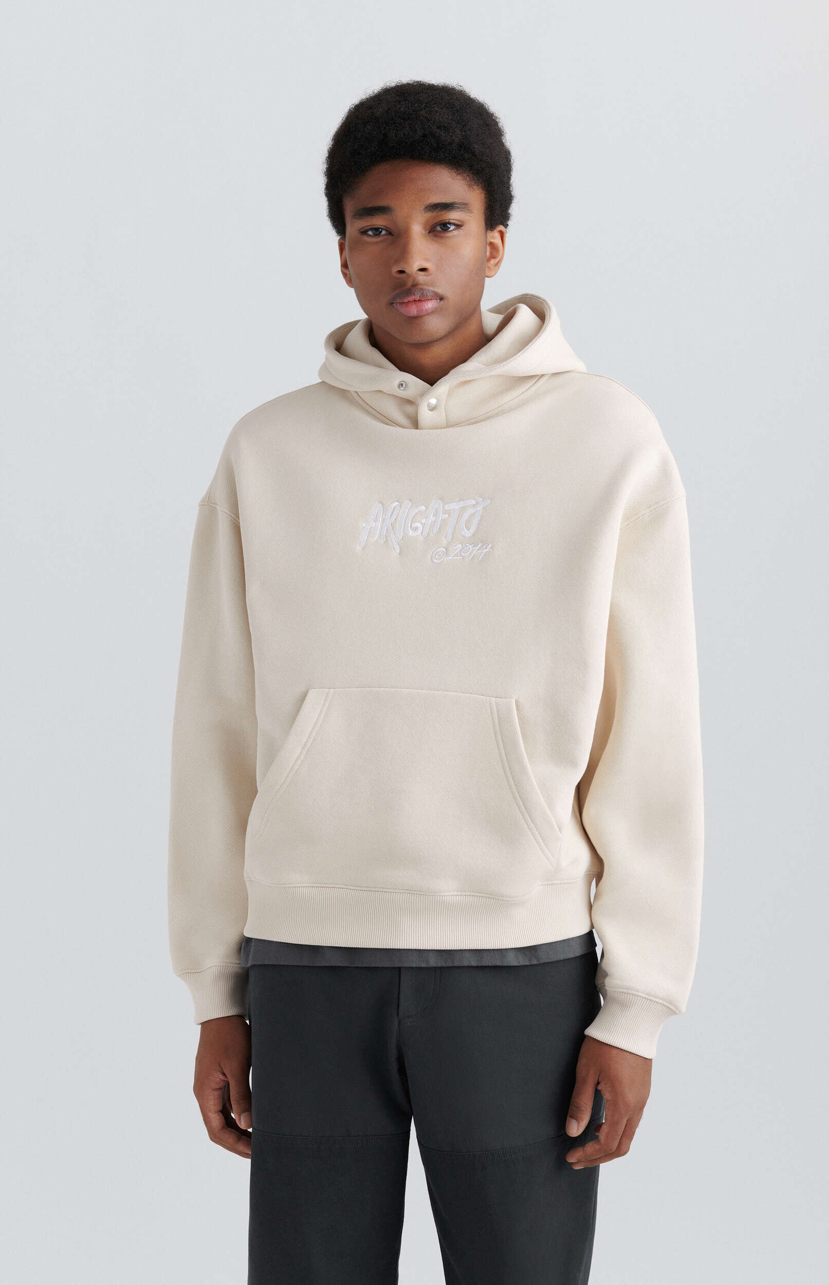 A young man with dark curly hair and a neutral expression stands facing forward, wearing a light beige hoodie with 'ARIGATO ©2014' embroidered on the chest. He is also wearing dark gray pants. The background is a plain light gray.