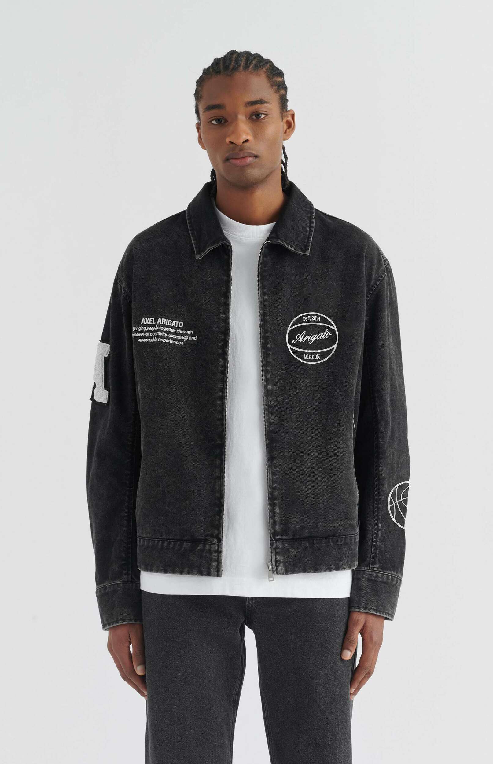 A young Black man with braided hair stands full-body against a plain white background, looking directly at the viewer. He is wearing a dark charcoal or washed black denim jacket with a zipper closure over a white t-shirt and dark wash jeans. The jacket has white embroidered text on the left chest that reads 'AXEL ARIGATO Bringing people together through power of positivity, community and memorable experiences' and a circular white logo on the right chest with 'EST. 2014 Arigato LONDON' inside. There are also white graphic details on the sleeves.