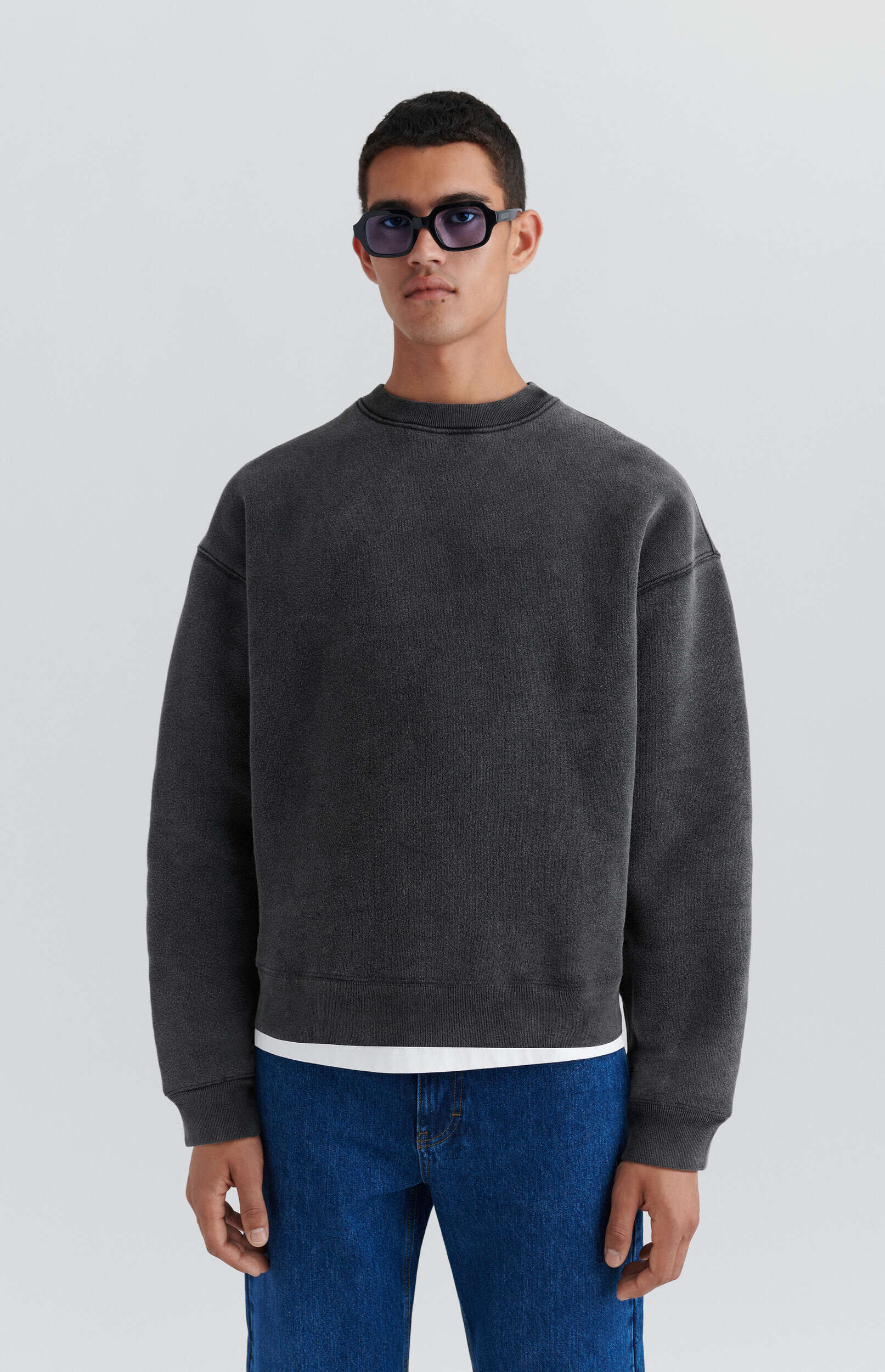 A man with short dark hair and angular black sunglasses stands against a plain white background, wearing a dark gray distressed crewneck sweatshirt over a white t-shirt, and blue jeans.