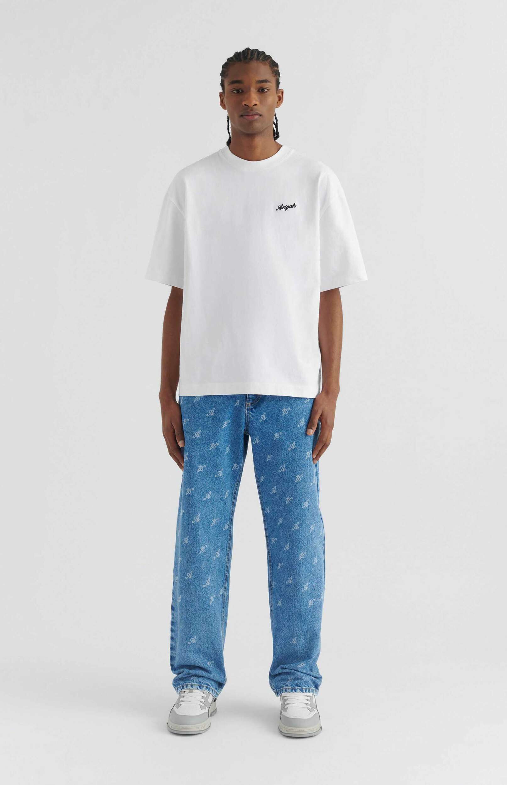 A young man with braided hair stands facing the camera against a plain white background. He is wearing a white short-sleeved t-shirt with 'Arigato' written in black script on the left chest, light blue jeans with a repeating white 'A' pattern, and light grey and white sneakers.