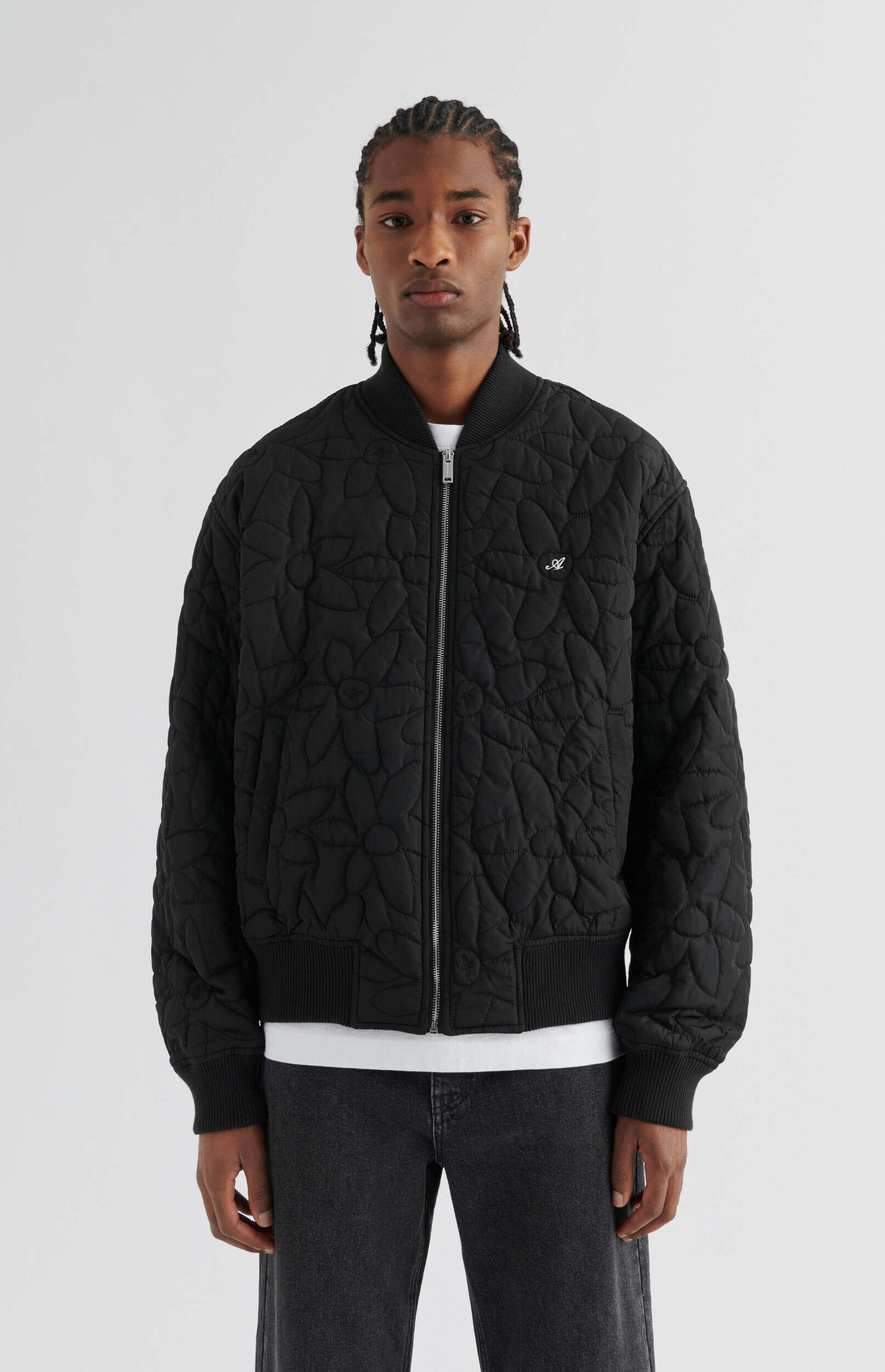 A young man with braided hair, wearing a black quilted bomber jacket with a subtle floral or leaf pattern, a white t-shirt, and dark jeans, stands facing forward against a plain white background.