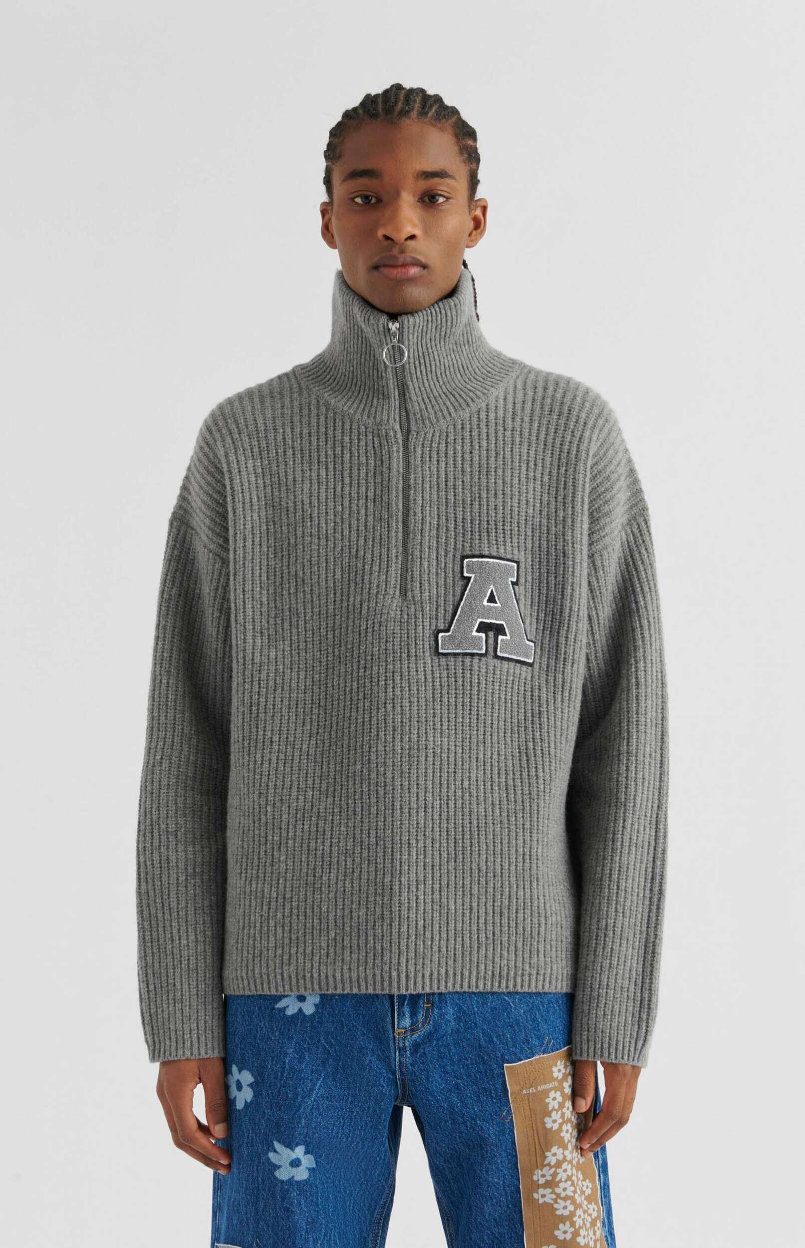 A young man with braided hair stands against a white background, wearing a grey ribbed knit turtleneck sweater with a quarter-zip. The sweater features a large grey 'A' patch with a black border on the left chest. He is also wearing blue denim jeans with faded white floral patterns and a light brown patch on the right thigh that also has a white floral print. He looks directly at the viewer.