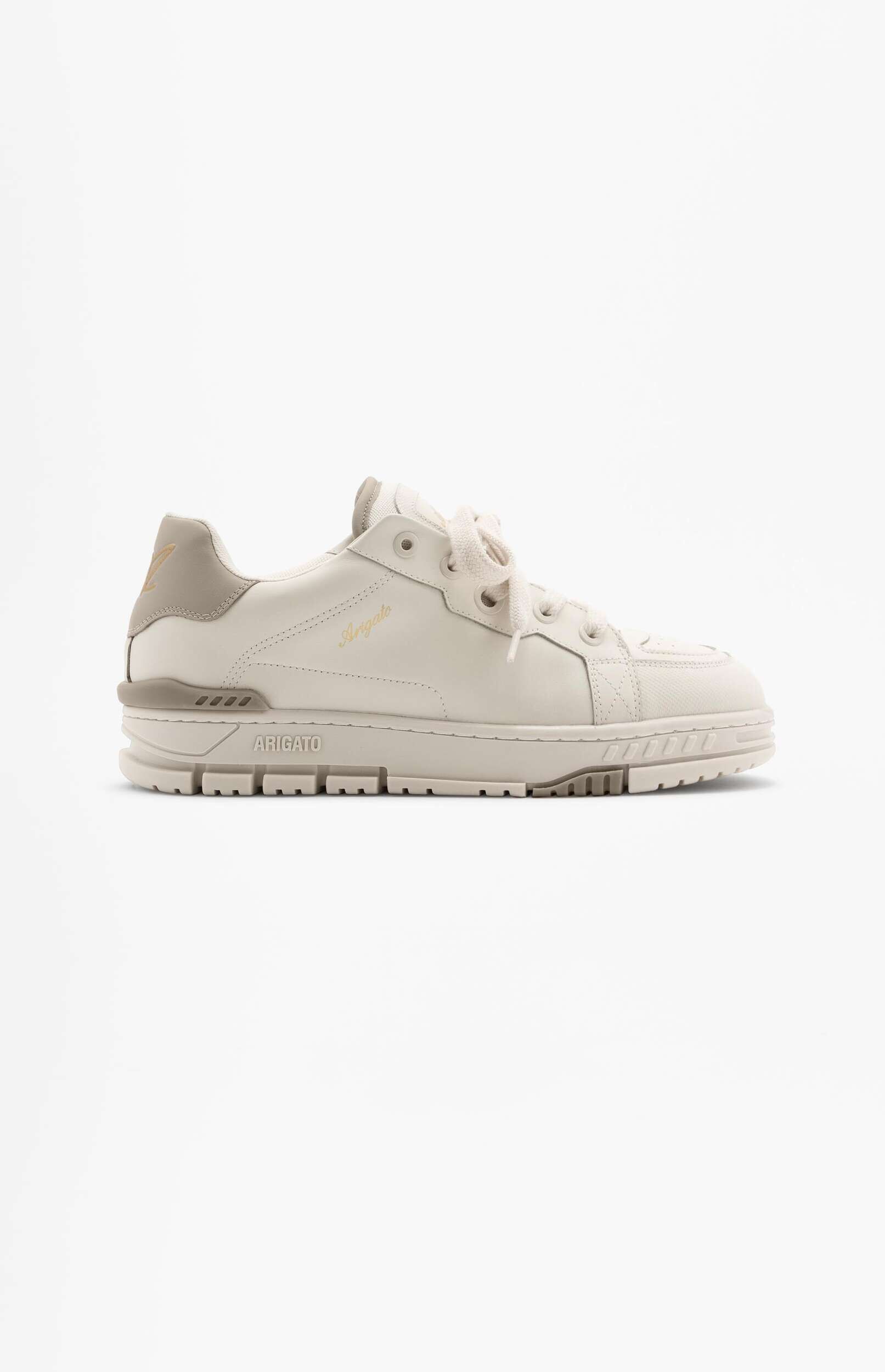 A side view of a cream or off-white sneaker with light beige accents, featuring a thick textured sole and "Arigato" branding in gold script on the side, set against a plain white background.