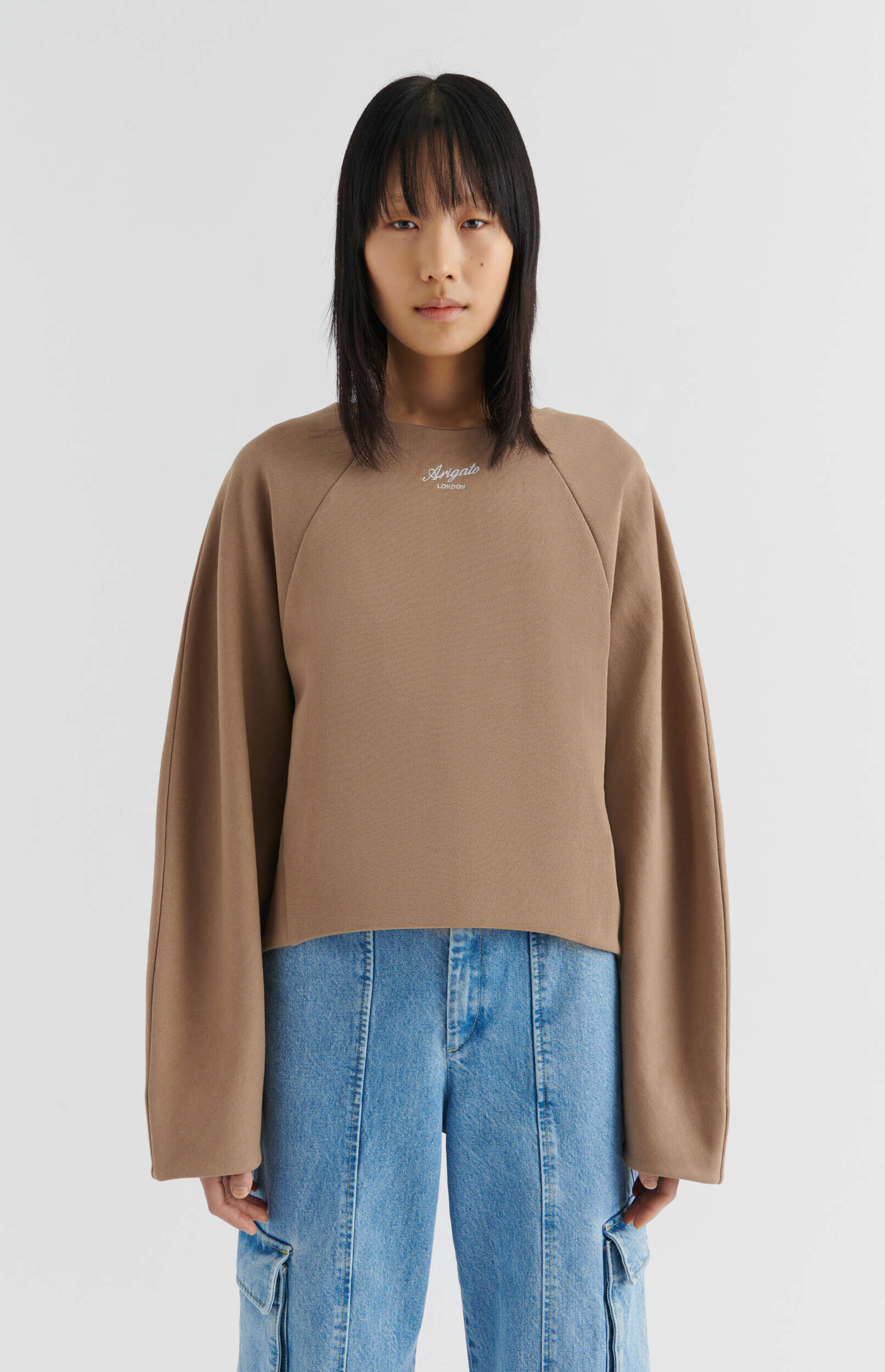 A person with straight black hair and bangs stands against a white background, wearing a brown crewneck sweatshirt with 'Arigato LONDON' embroidered on the front and light blue denim cargo jeans.