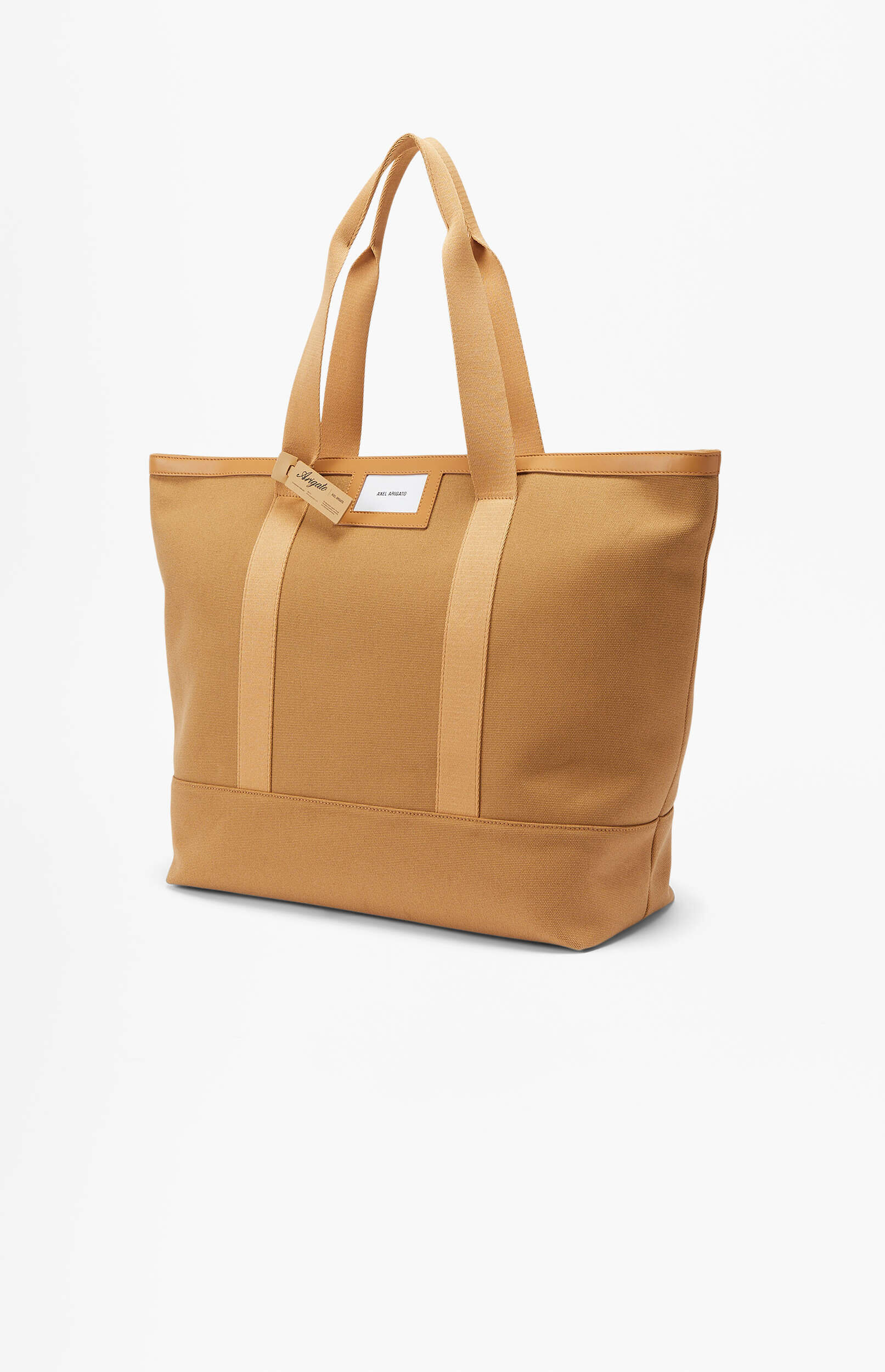 A light brown canvas tote bag with wide straps and leather trim, featuring a white rectangular patch with 'AXEL ARIGATO' on the front and a smaller 'Arigato' tag hanging from a strap, set against a plain white background.