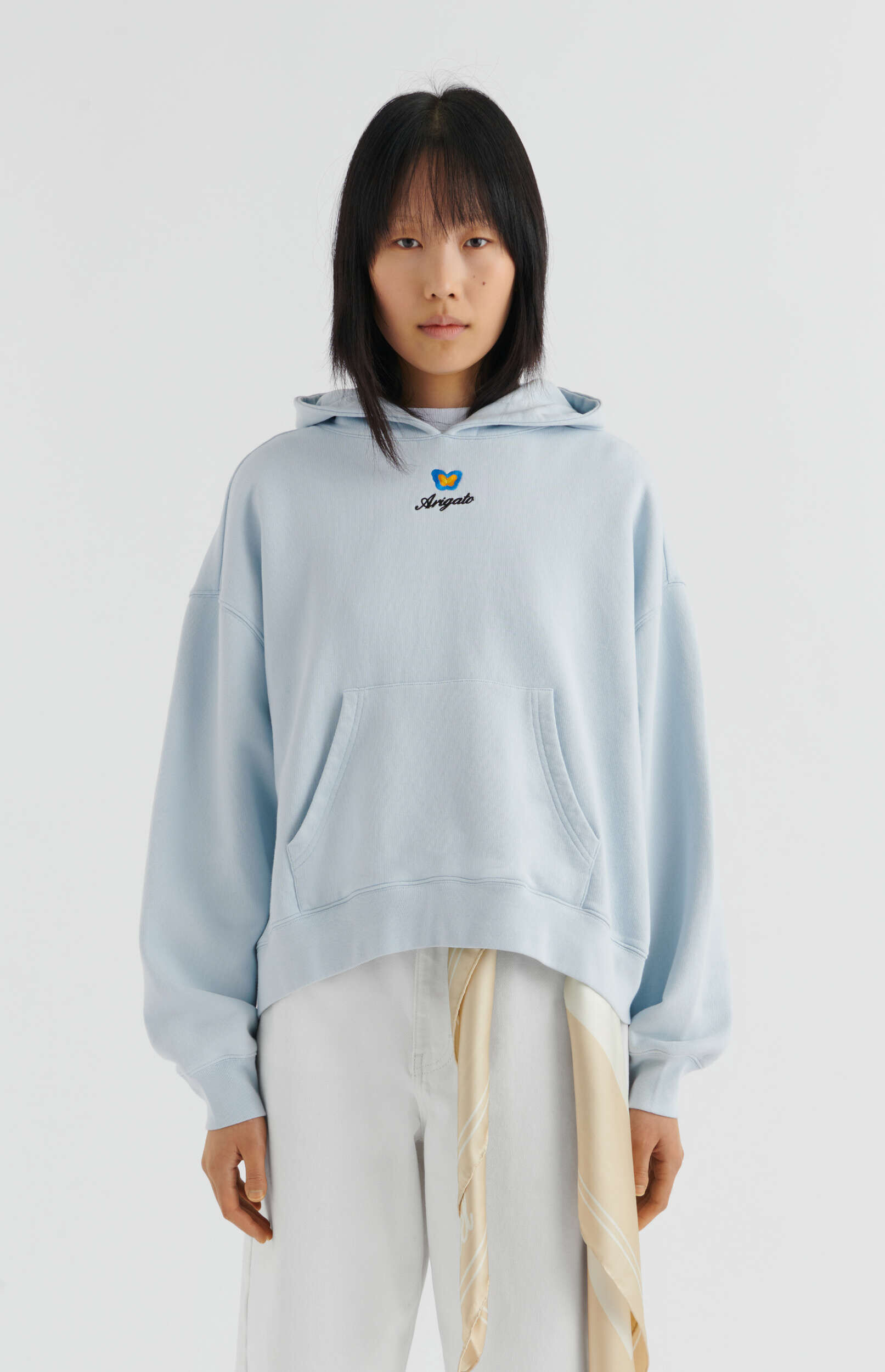 A young woman with short black hair and bangs stands against a white background, looking directly forward. She wears a light blue oversized hooded sweatshirt with a small black 'Arigato' text and a yellow and blue butterfly logo embroidered on the chest. She is also wearing white trousers, and a light beige and white striped silk scarf is draped over her right hip.