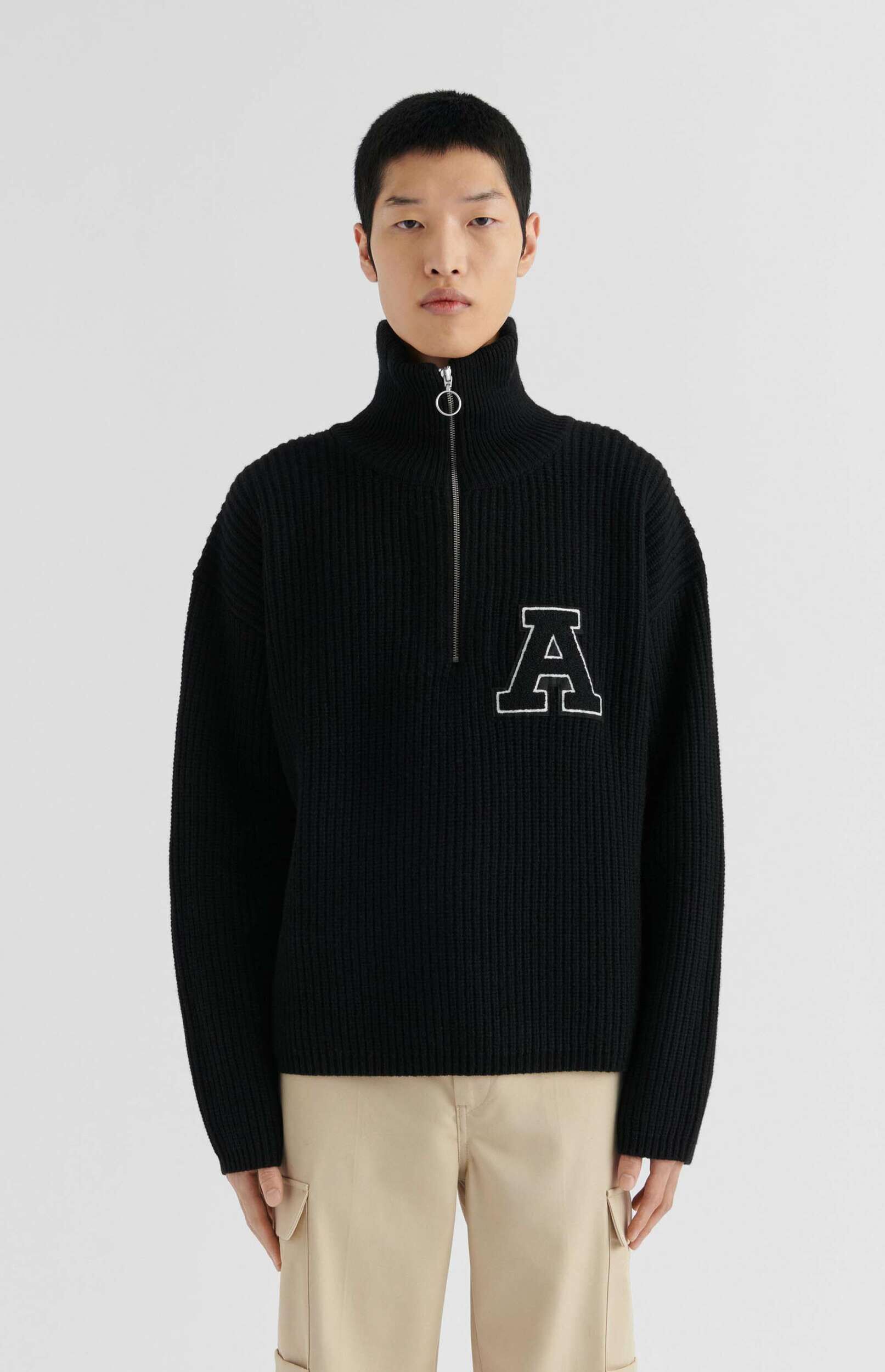 A person wears a black ribbed half-zip turtleneck sweater featuring a prominent white 'A' patch on the chest, paired with light beige cargo pants, standing against a plain white background.