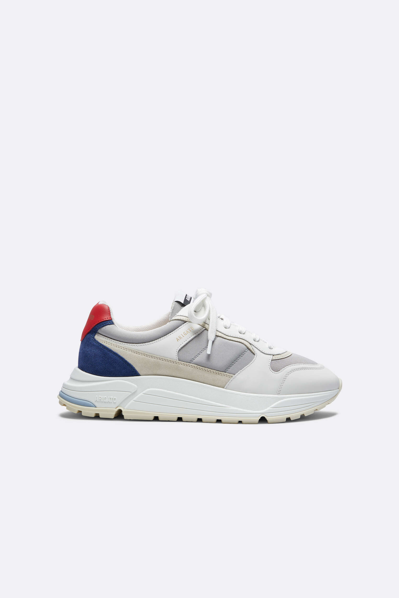 A single Axel Arigato sneaker displayed from a side view against a white background. The shoe features a multi-panel design in shades of white, light grey mesh, beige suede, and dark blue suede on the heel, topped with a striking red leather heel tab. It has white laces and a chunky off-white sole with a subtle light blue detail near the rear.