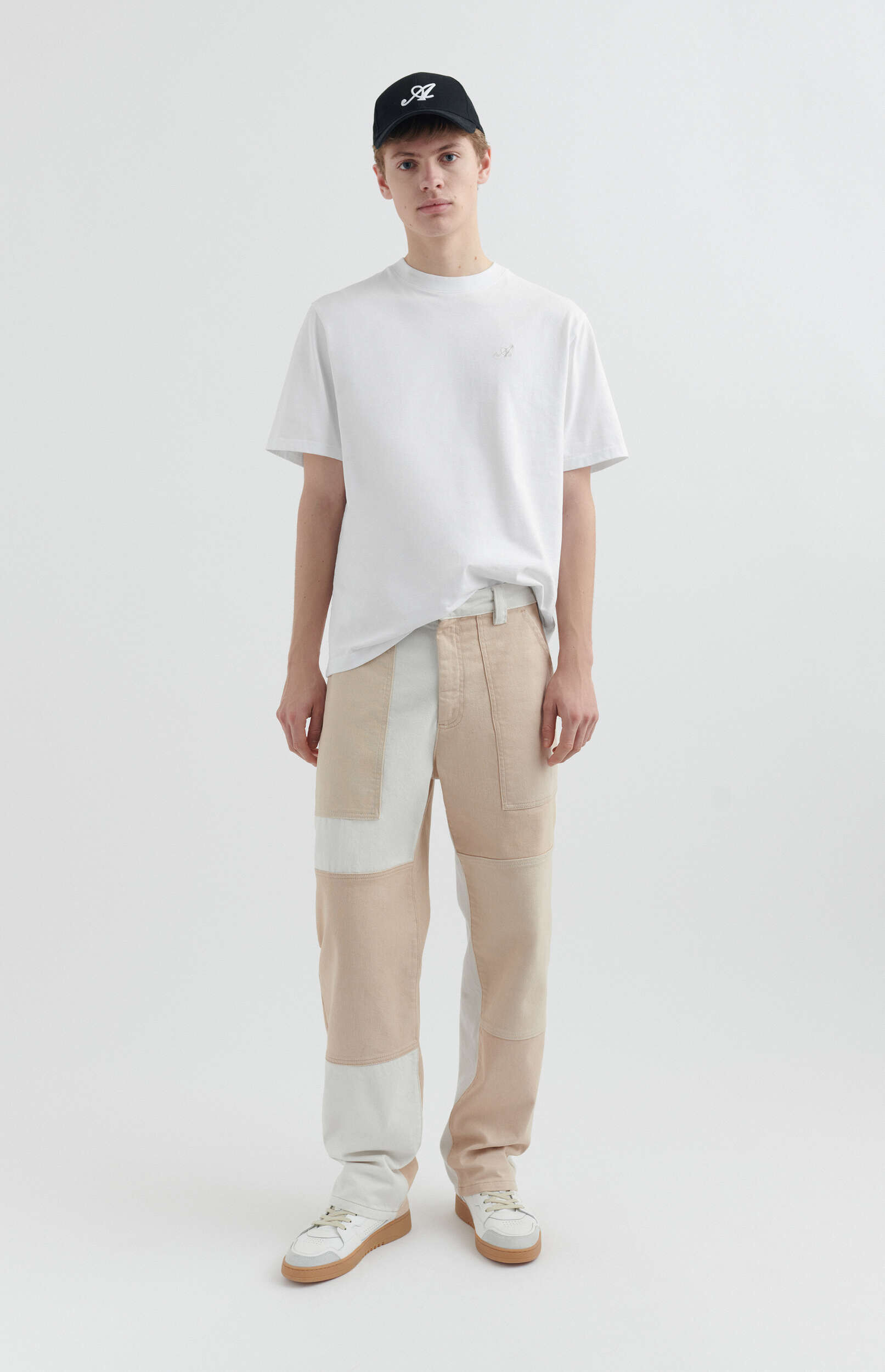 A young man stands full-length against a white background, wearing a white crew neck t-shirt with a small embroidered logo, a black baseball cap with a white script logo, and unique patchwork pants in shades of light beige and off-white. He completes the look with white and grey sneakers featuring gum soles.